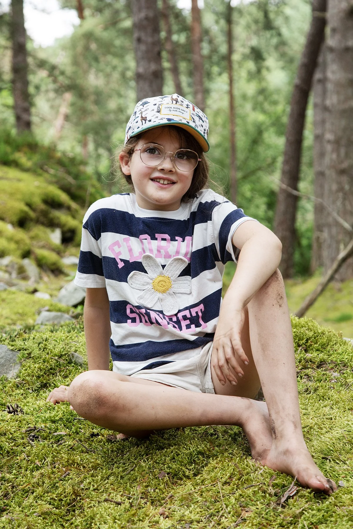A young girl with glasses, sitting on moss in a forest, wearing a striped t-shirt with a flower that says 'Florida Sunset', and a cap.