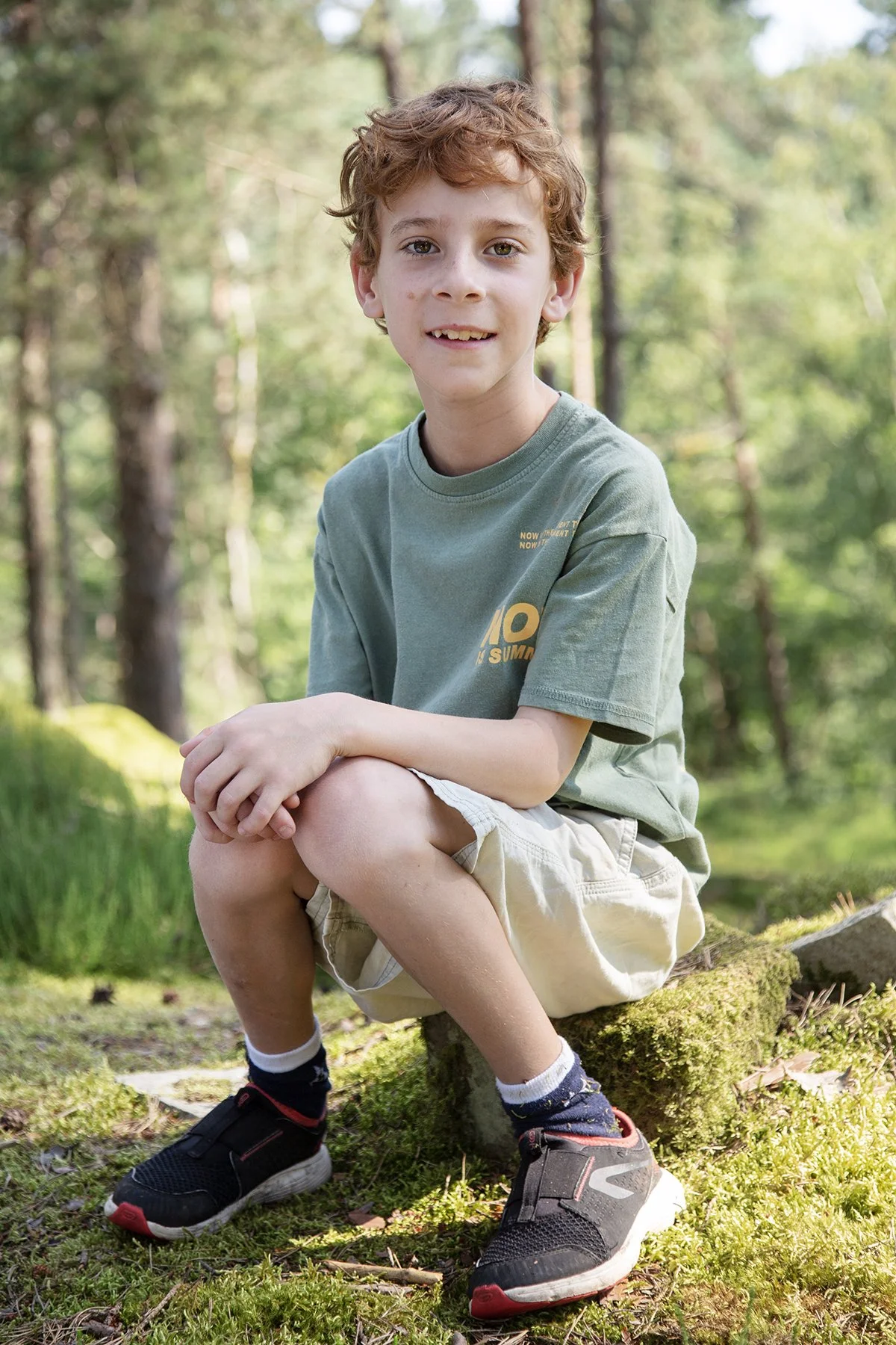 A young boy with curly red hair sitting on a moss-covered log in a forest of tall trees with green foliage.