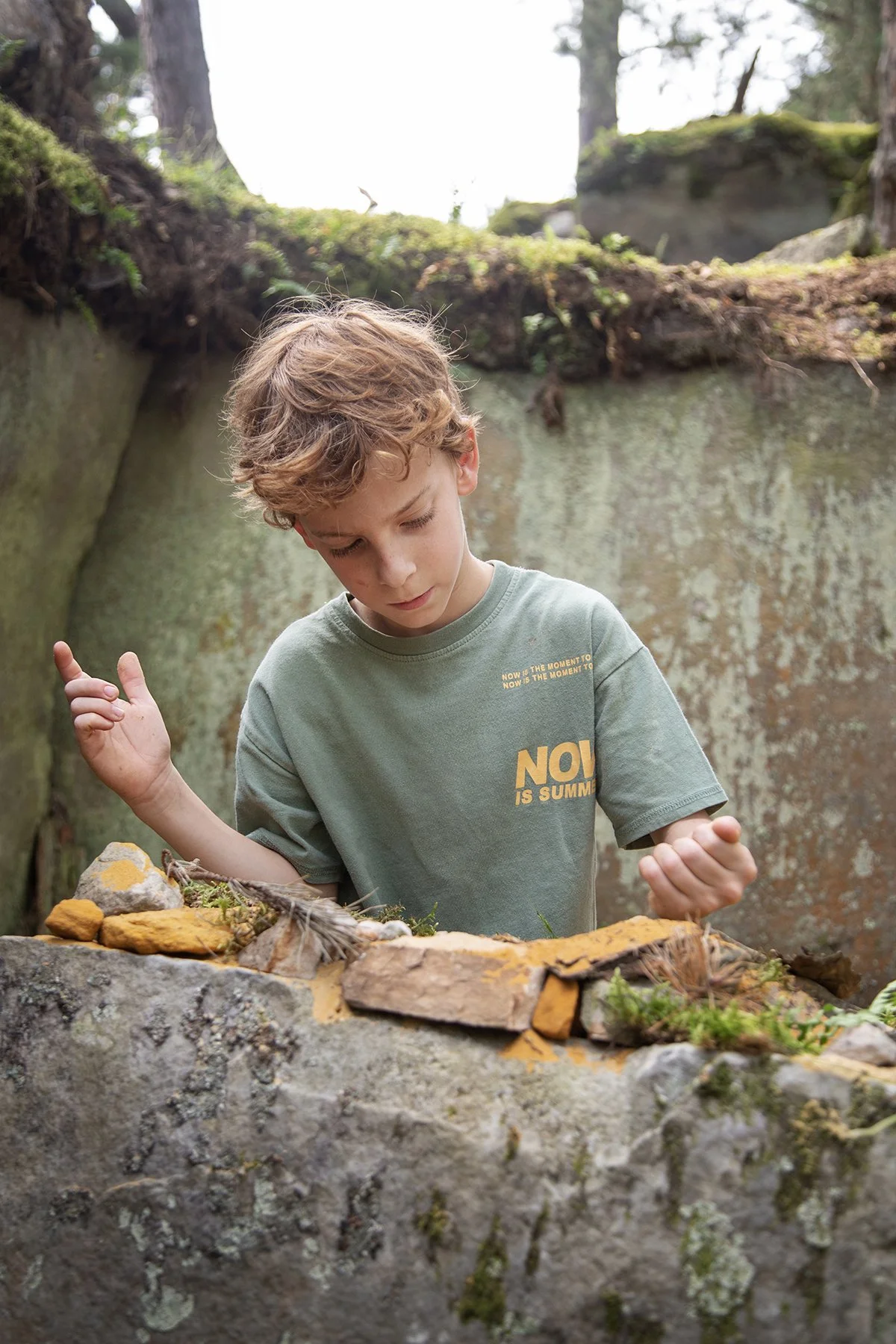 A young boy with curly blonde hair outdoors, standing behind a large moss-covered stone wall, surrounded by trees, looking down at objects on the wall with a thoughtful expression.
