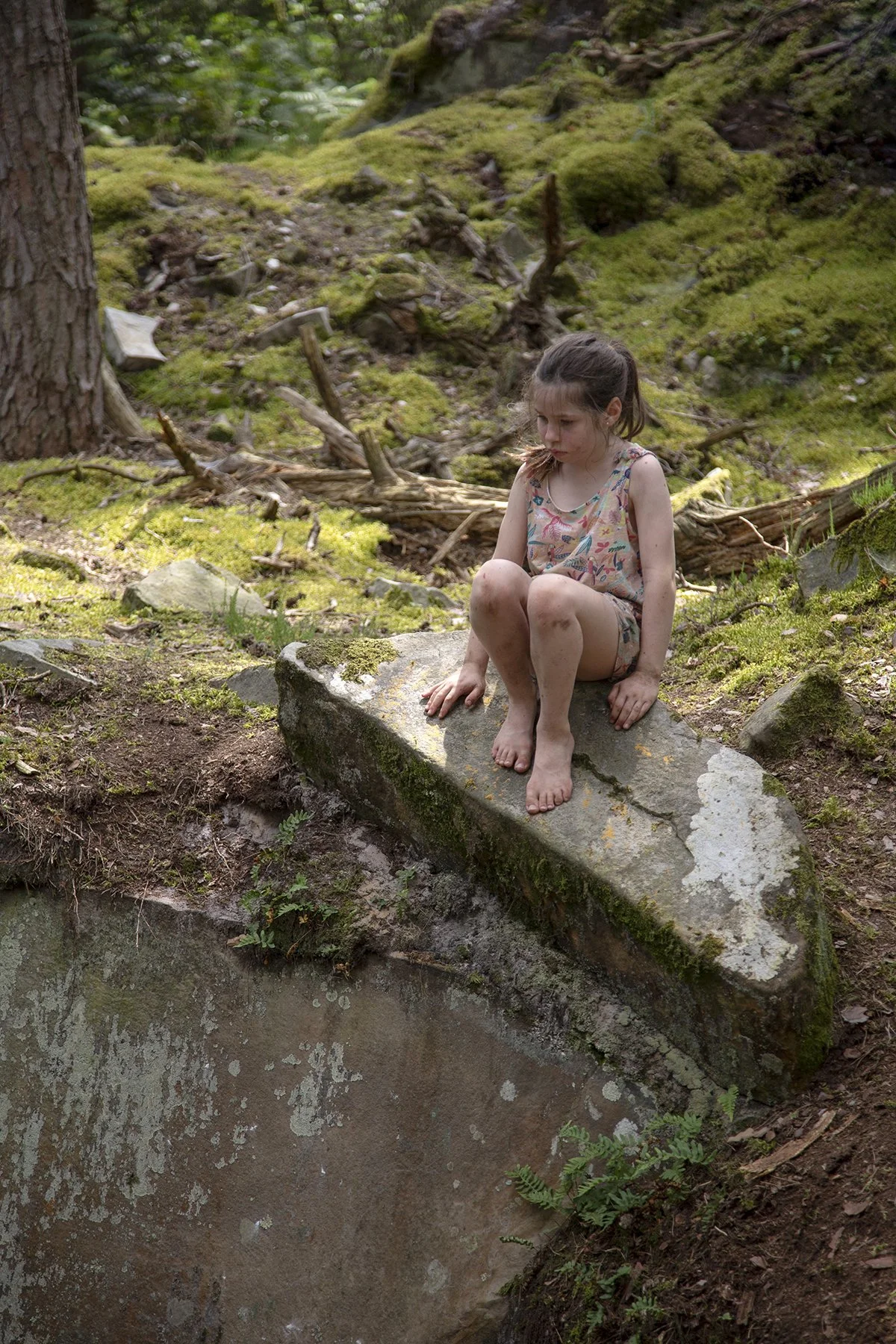 A young girl with brown hair in a ponytail sits on a moss-covered rock in a forest, looking down thoughtfully. She is wearing a sleeveless, colorful dress and is barefoot. The background consists of green moss, fallen branches, and trees, with dapple