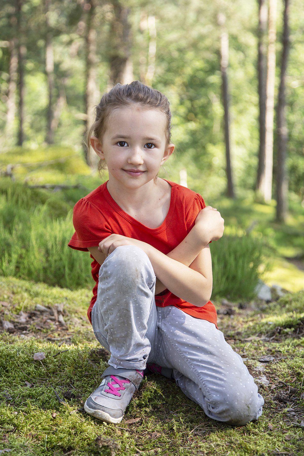 A young girl with a red shirt and gray pants kneeling on moss and grass in a lush forest, smiling at the camera.