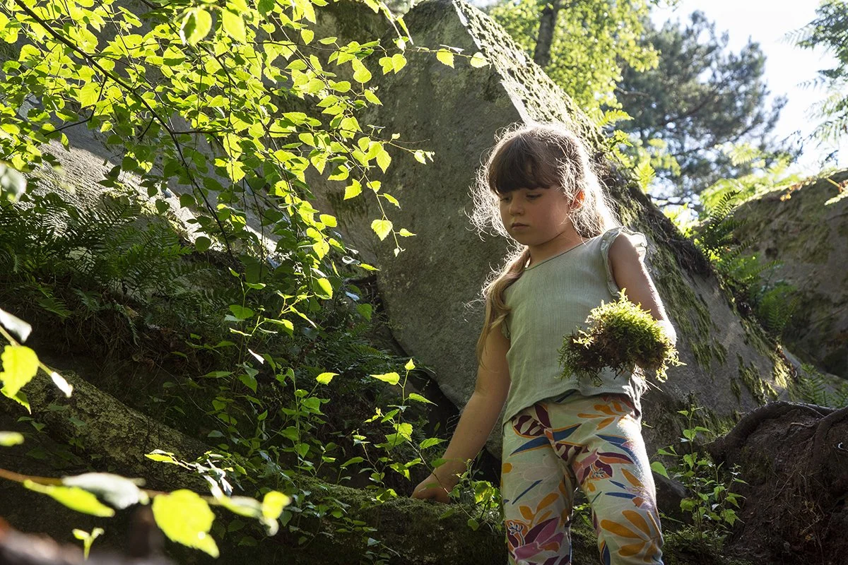 Young girl in a sleeveless top and colorful patterned pants standing outdoors in a forested area, holding a bunch of moss, surrounded by green foliage and rocks.