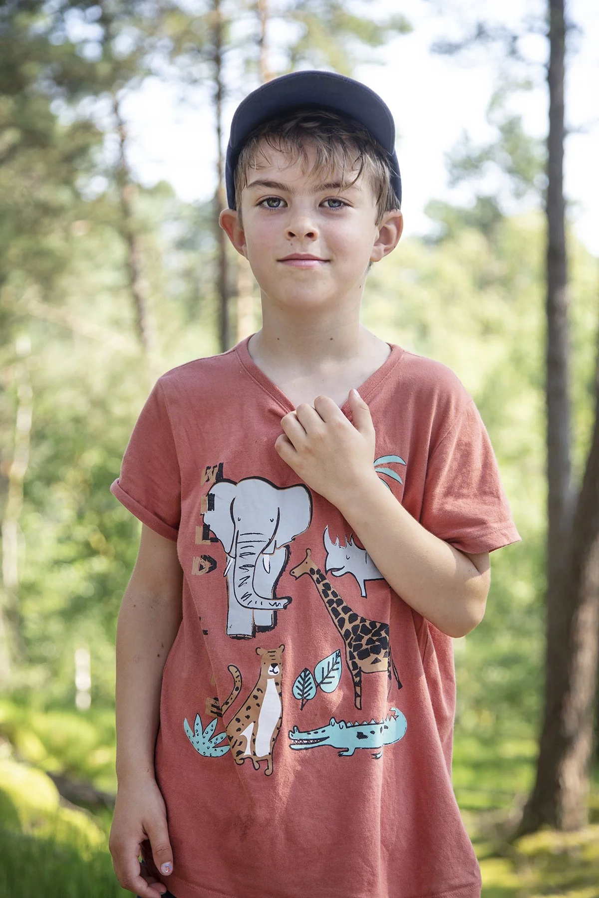 A young boy with short light brown hair wearing a dark baseball cap and a pink T-shirt with animal illustrations, standing outdoors in a wooded area with trees in the background.