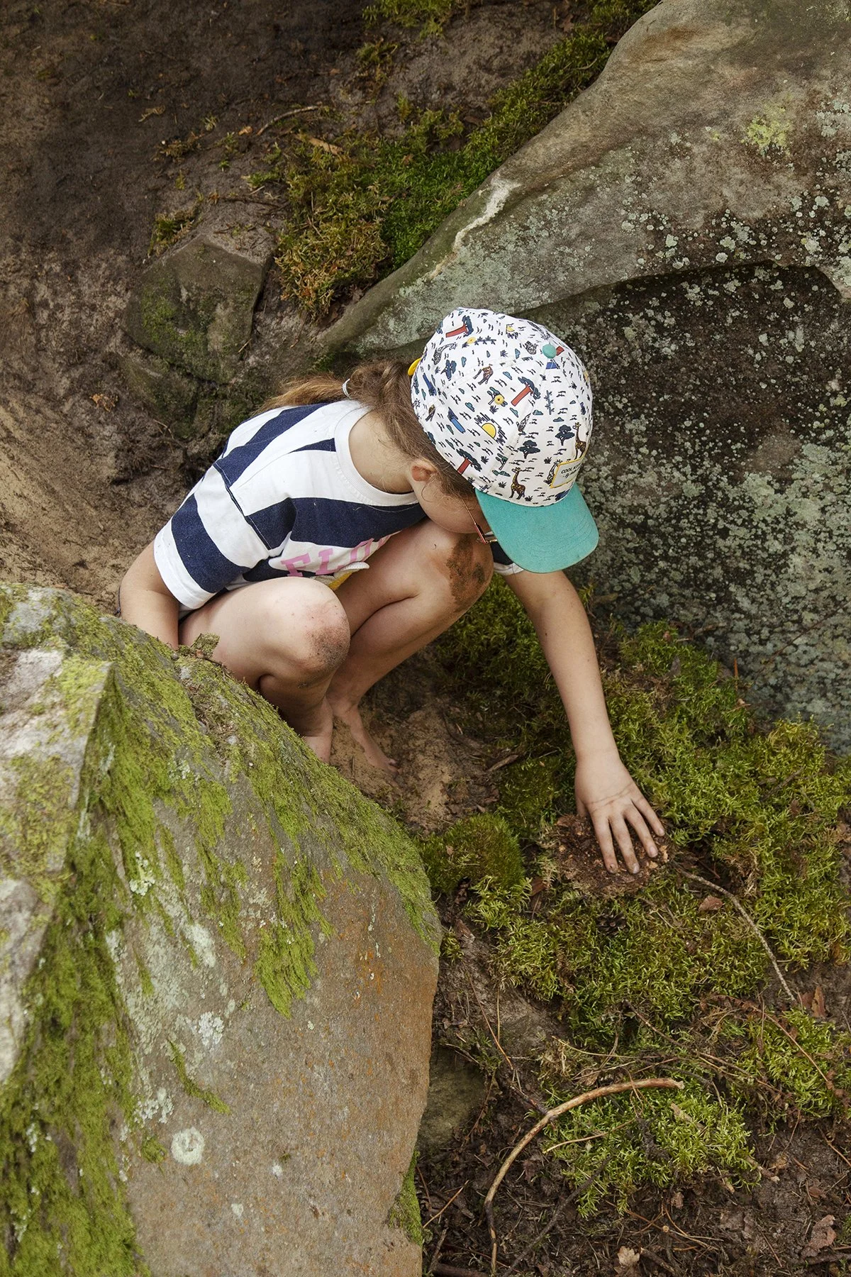 Child exploring outdoors, crouched near moss-covered rocks, wearing a colorful patterned hat, glasses, and a striped shirt.