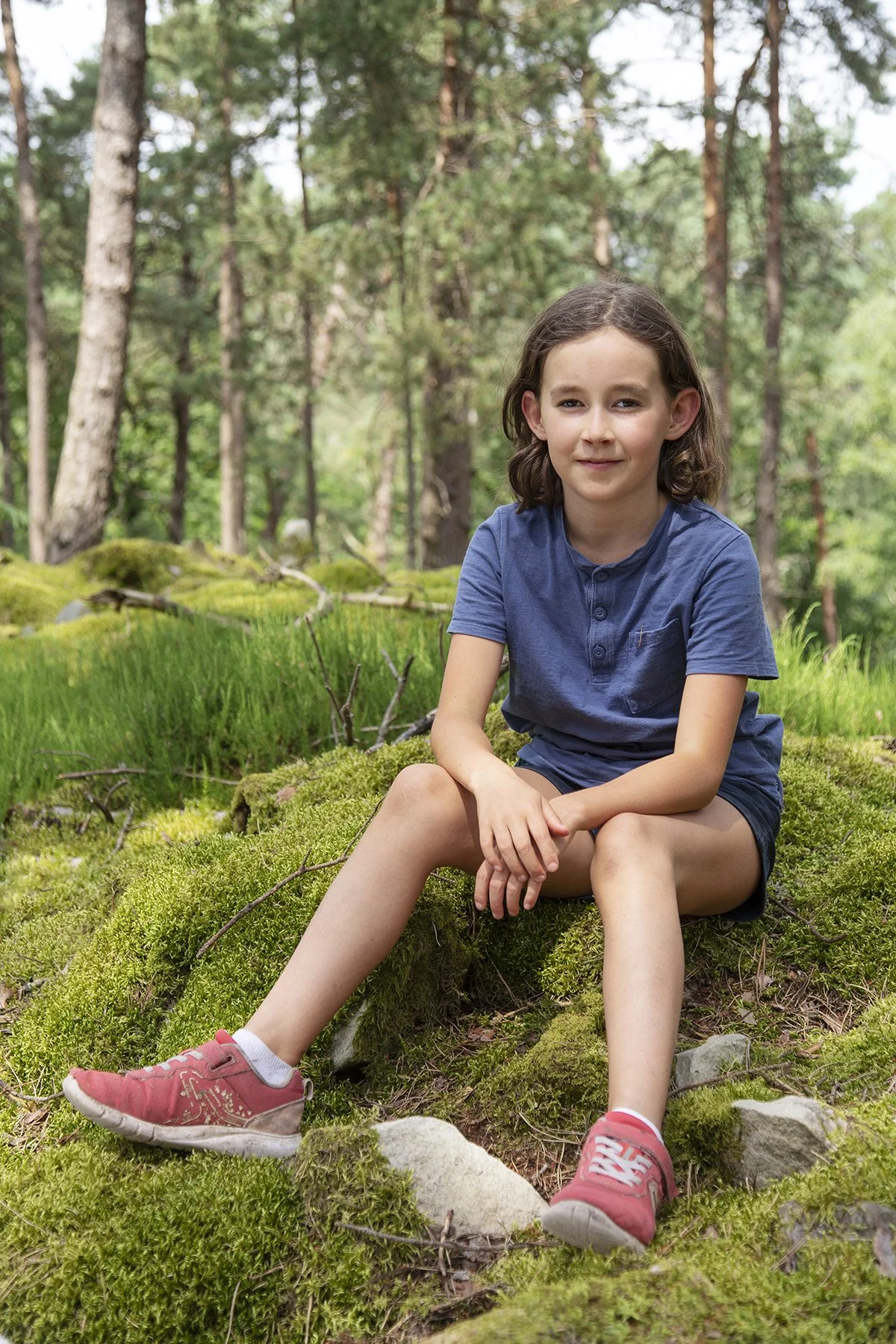 A young girl sitting on moss in a forest, wearing a blue shirt, dark shorts, and pink sneakers, with trees in the background.