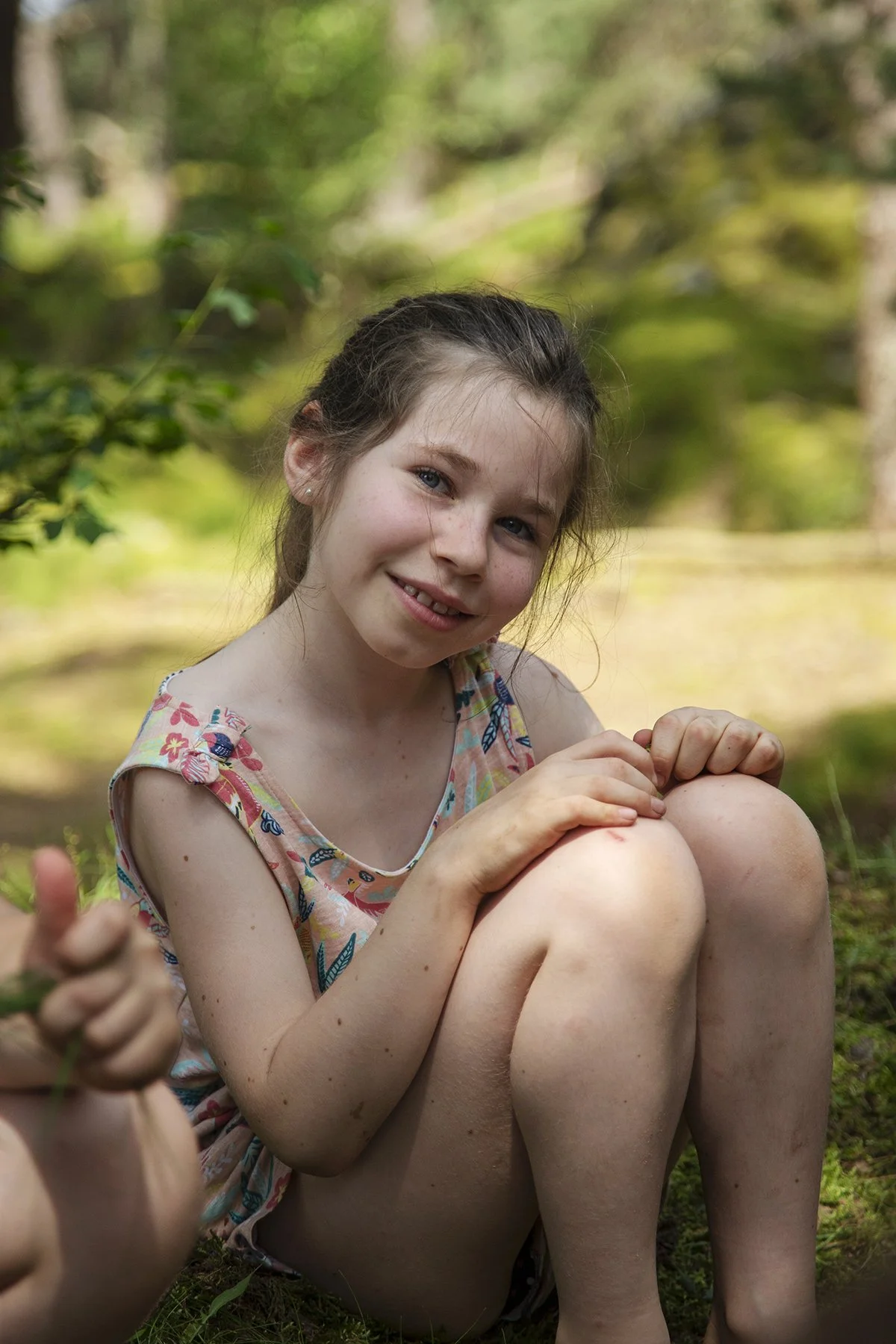 A young girl sitting outdoors on grass with her knees hugged to her chest, smiling at the camera. She is wearing a sleeveless top with a colorful floral pattern, and her hair is tied back. The background shows a blurred green wooded area.