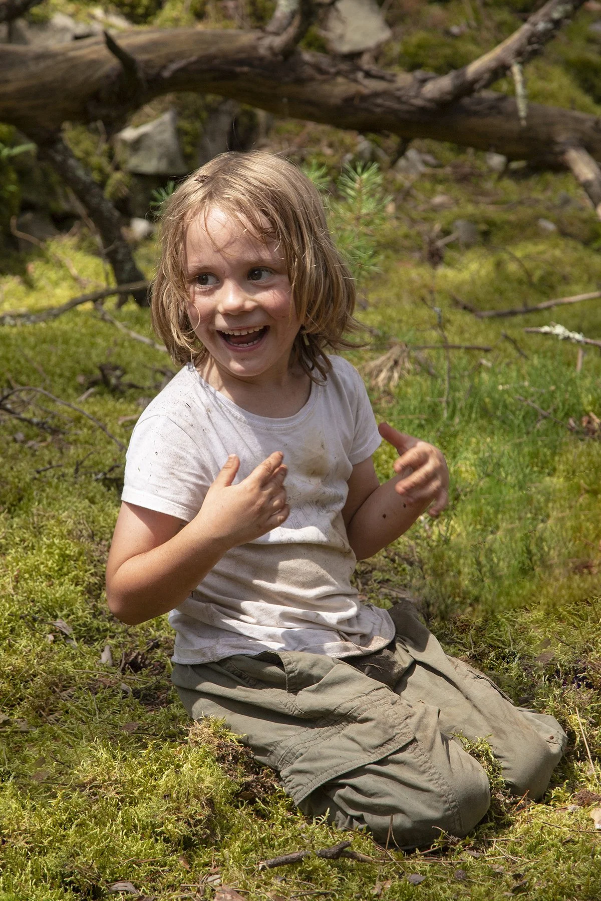 A young girl with wet, messy hair and a white t-shirt, sitting on the forest floor, smiling and pointing.