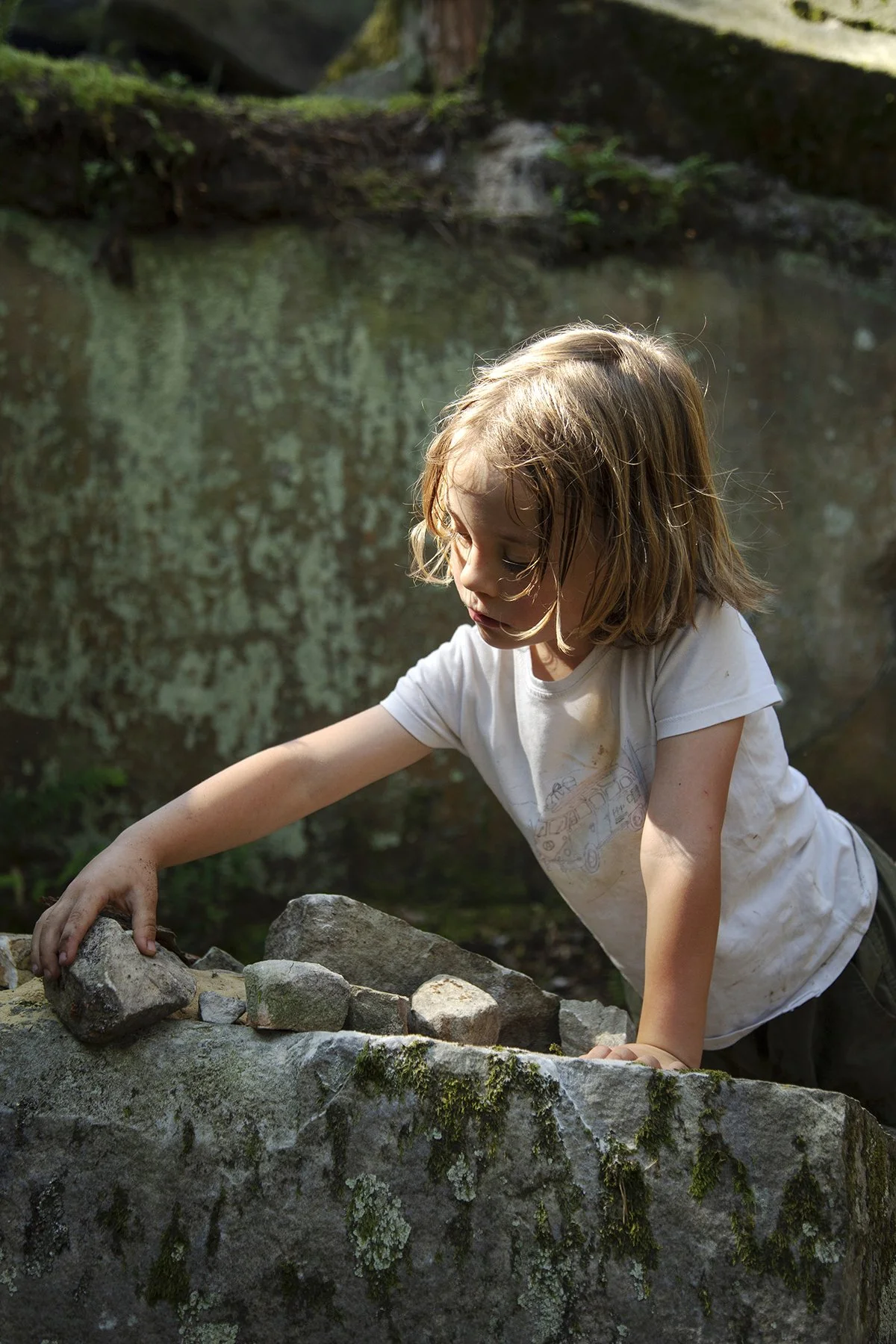 A young girl in a white t-shirt exploring outdoors, reaching out to touch rocks on a moss-covered stone in a natural setting.