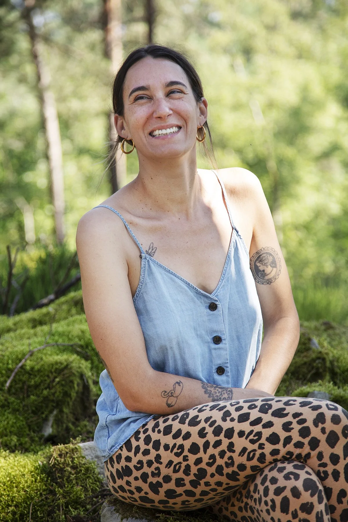 A woman sitting outdoors on mossy ground surrounded by trees, wearing a blue sleeveless top and leopard print leggings, smiling and looking away
