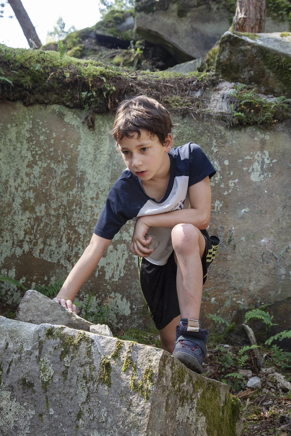 A young boy climbing over a large moss-covered rock outdoors in a forested area.
