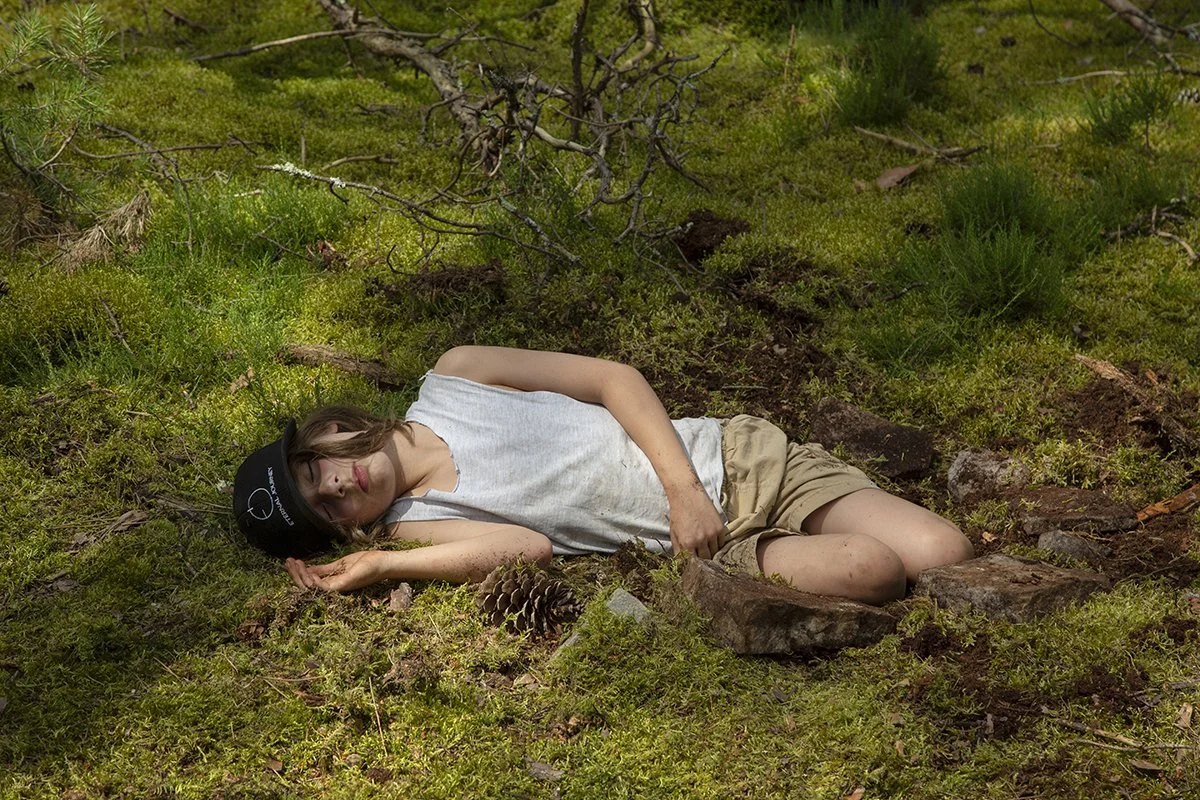 A young woman lies on the forest floor among greenery and rocks, with her eyes closed and arm under her head, wearing a sleeveless white shirt, tan shorts, and a black cap.