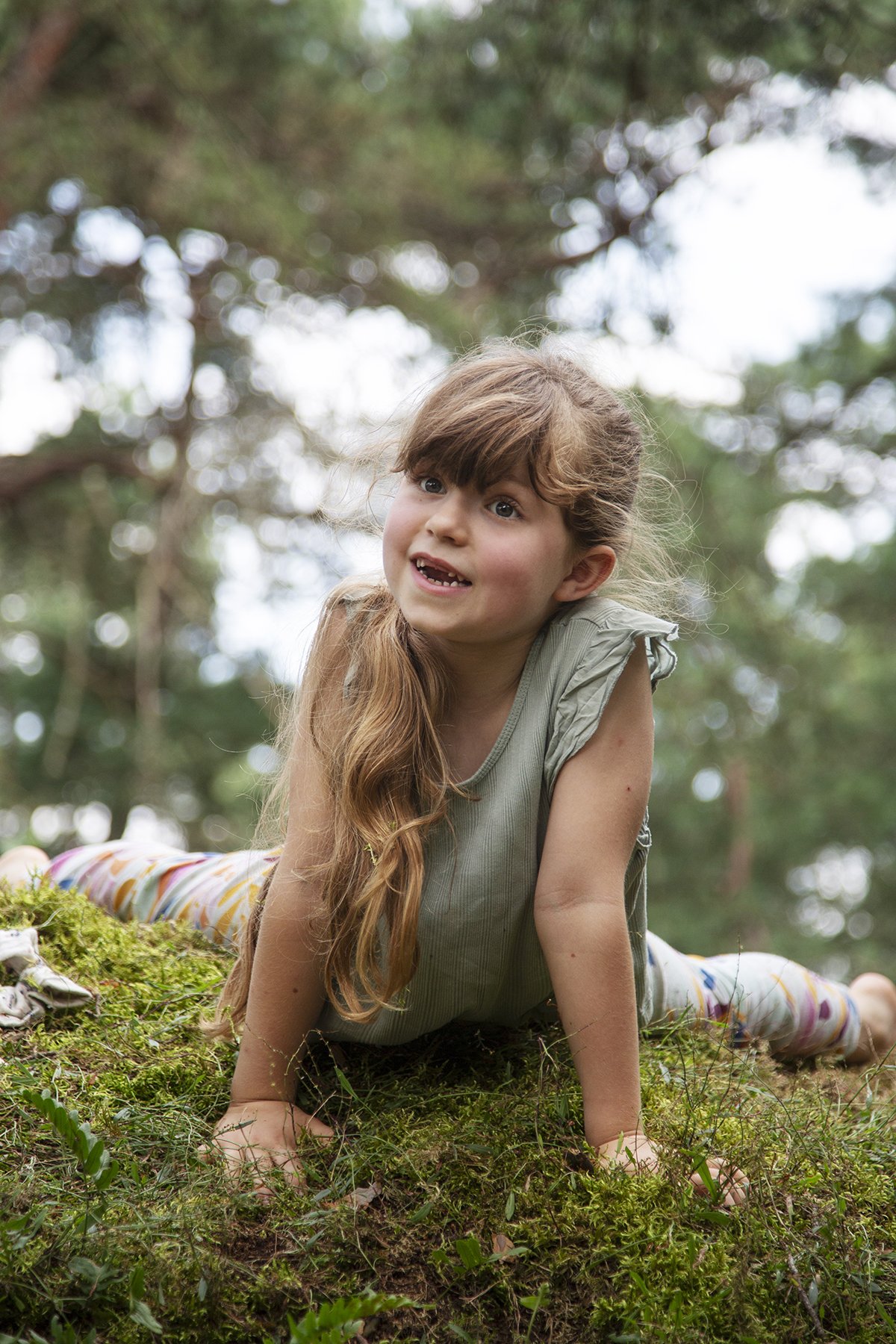 Young girl with long hair, lying on her stomach on green mossy ground in a forest, smiling and looking up, surrounded by tall trees and blurred leafy background.