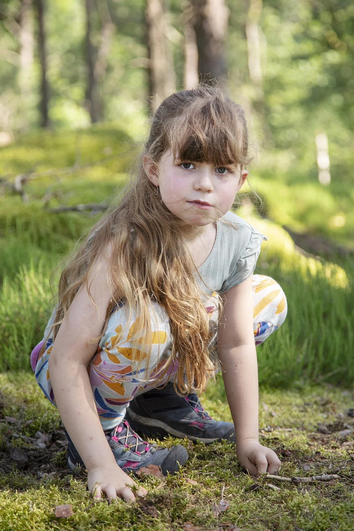 A young girl with long brown hair and blue eyes crouching on grass in a wooded area.