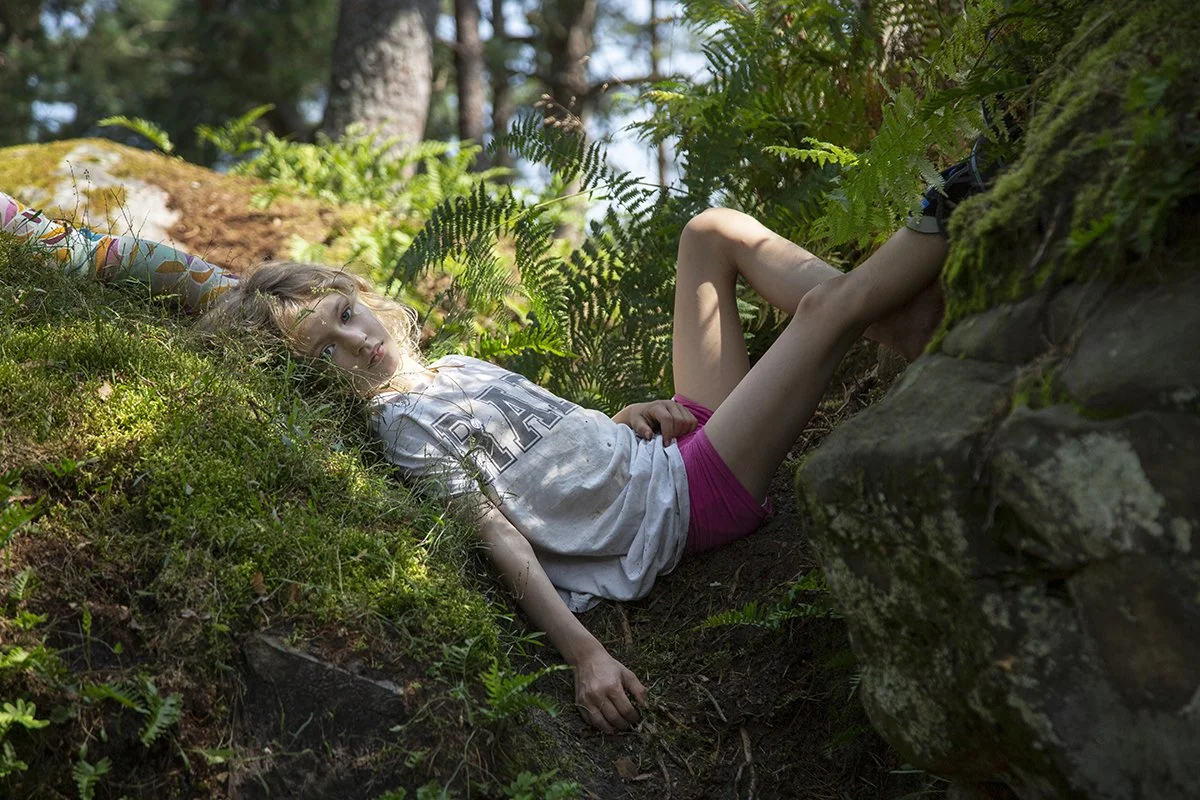 A young girl with blonde hair lies on the mossy ground in a forest, surrounded by green ferns and trees, wearing a white T-shirt and pink shorts, looking at the camera.