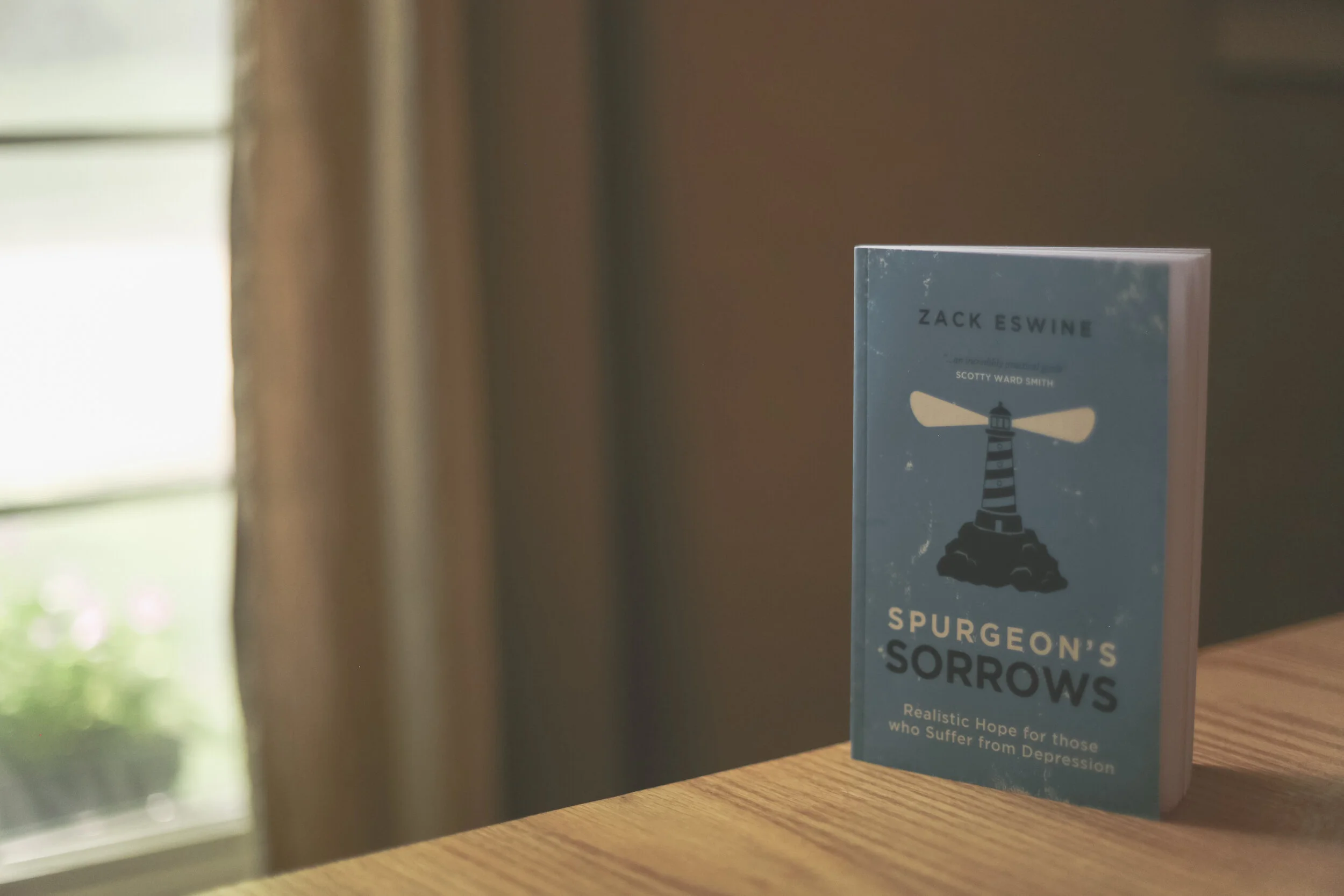 A book titled 'Spurgeon's Sorrows' by Zack Eswine, with a lighthouse illustration, sitting on a wooden surface near a window with a curtain.