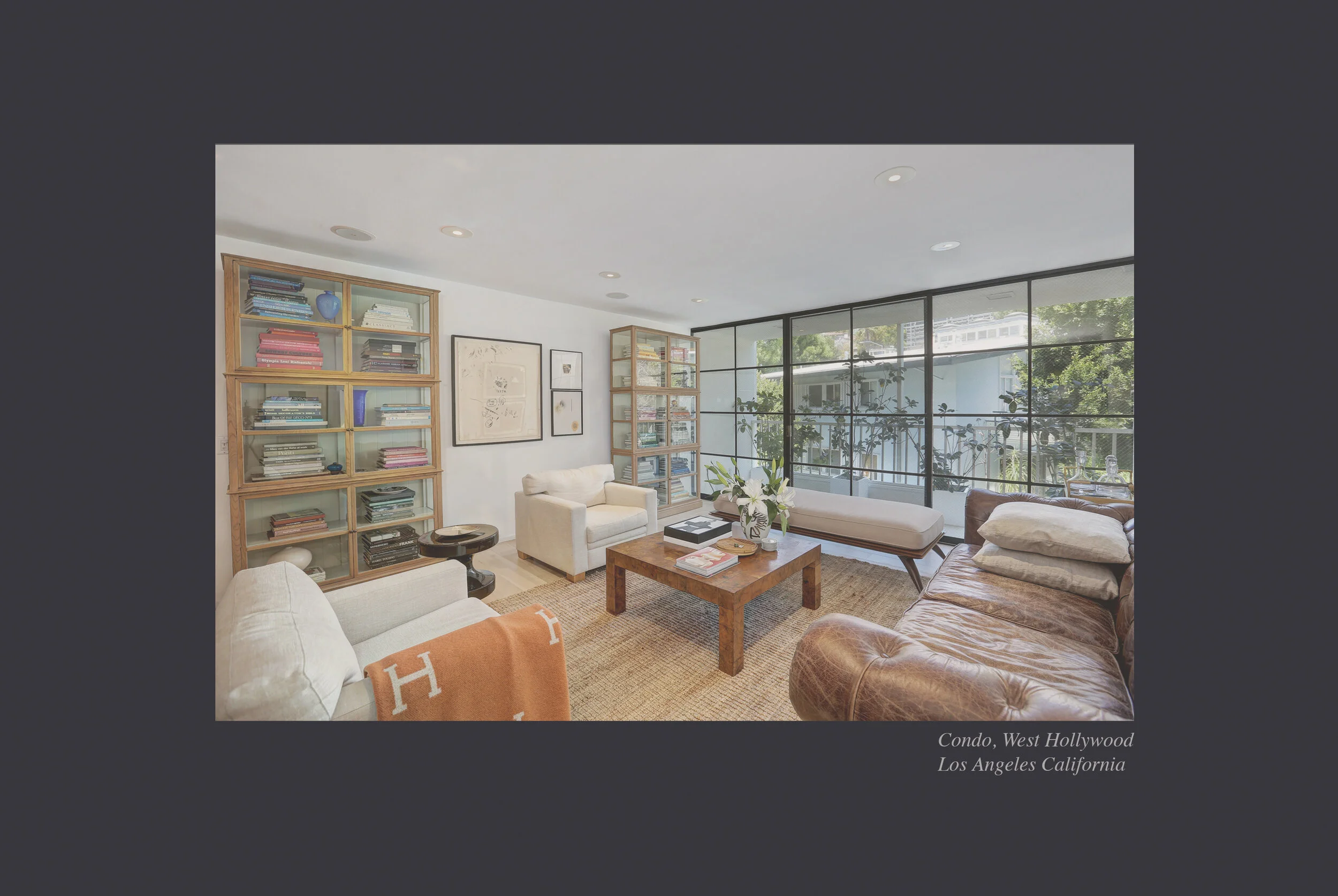 Living room with beige and brown sofas, wooden coffee table with books and a floral vase, floor-to-ceiling windows showing greenery outside, and built-in bookshelves with books and decorative items