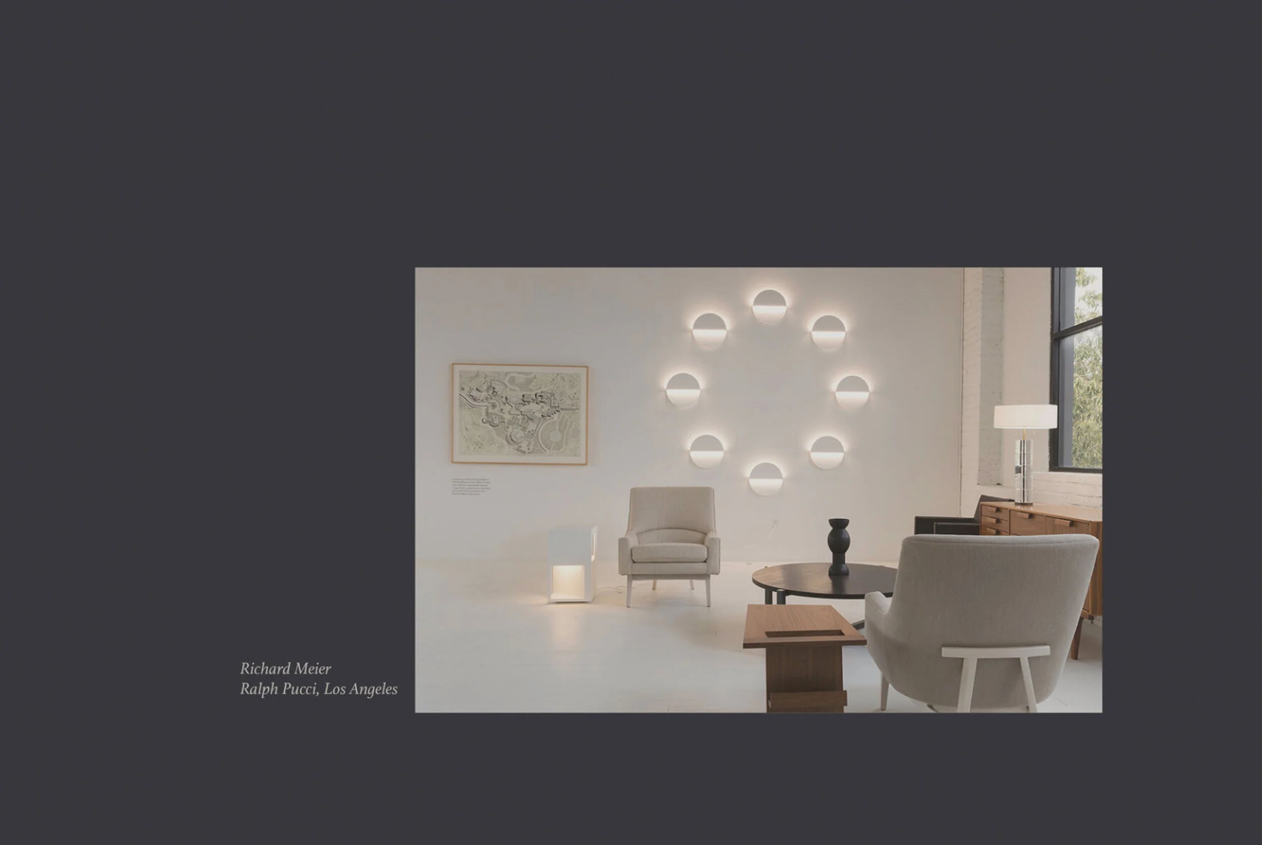 Interior of a modern, minimalist living room with white walls, a circular wall light installation, a framed map on the wall, a beige armchair, a round coffee table with black vases, a white floor lamp, and large windows with a view of greenery outsid