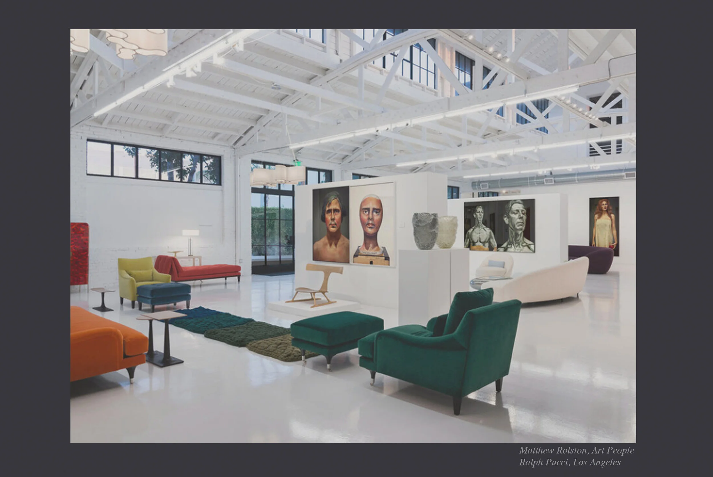 Interior of an art gallery with white walls and exposed ceiling beams, various colorful chairs, art sculptures, and portraits of people displayed on white partition walls.