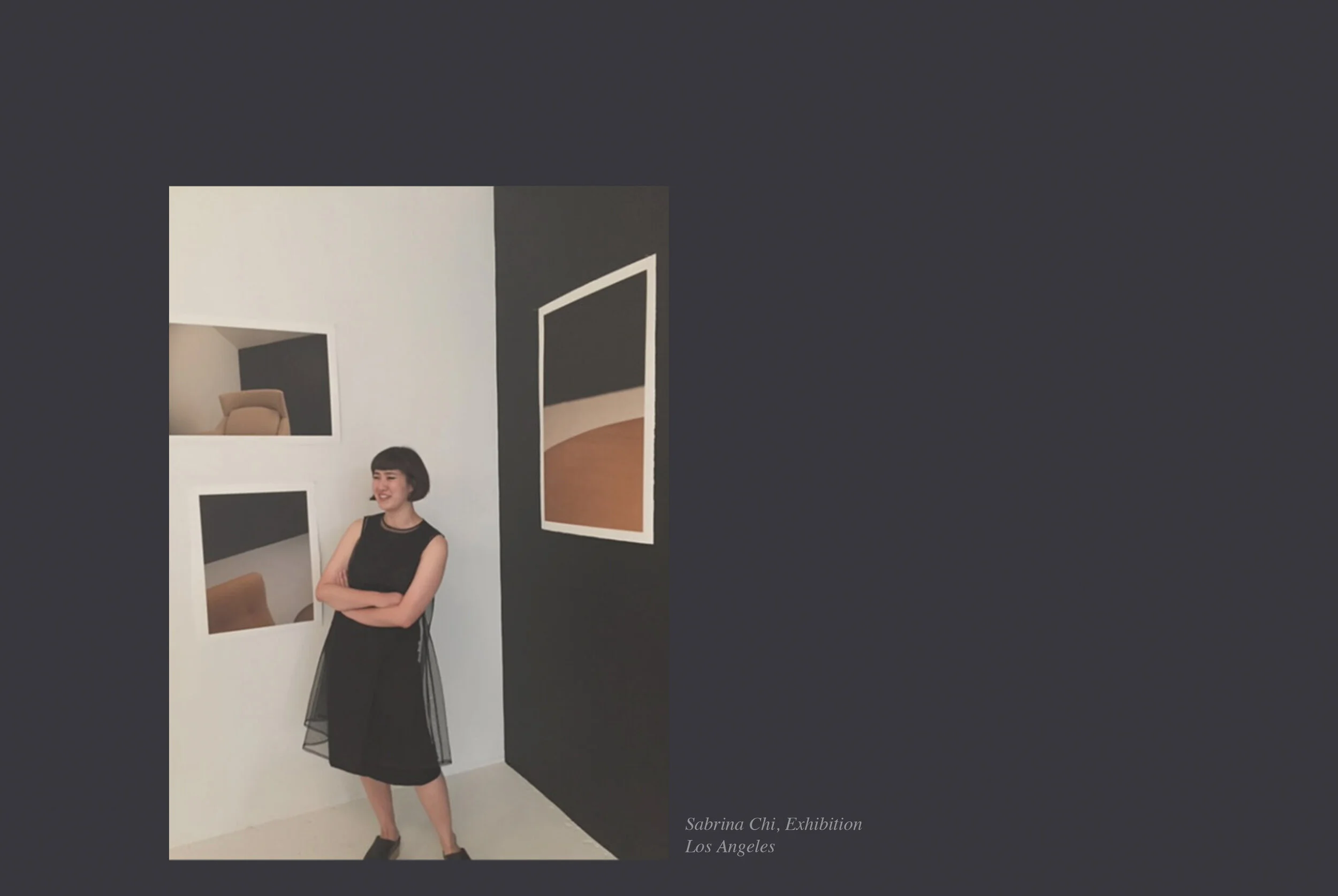 Photograph of a woman standing in front of a white wall with black and brown artwork, posing with crossed arms and wearing a black dress, at Sabrina Chi's exhibition in Los Angeles.