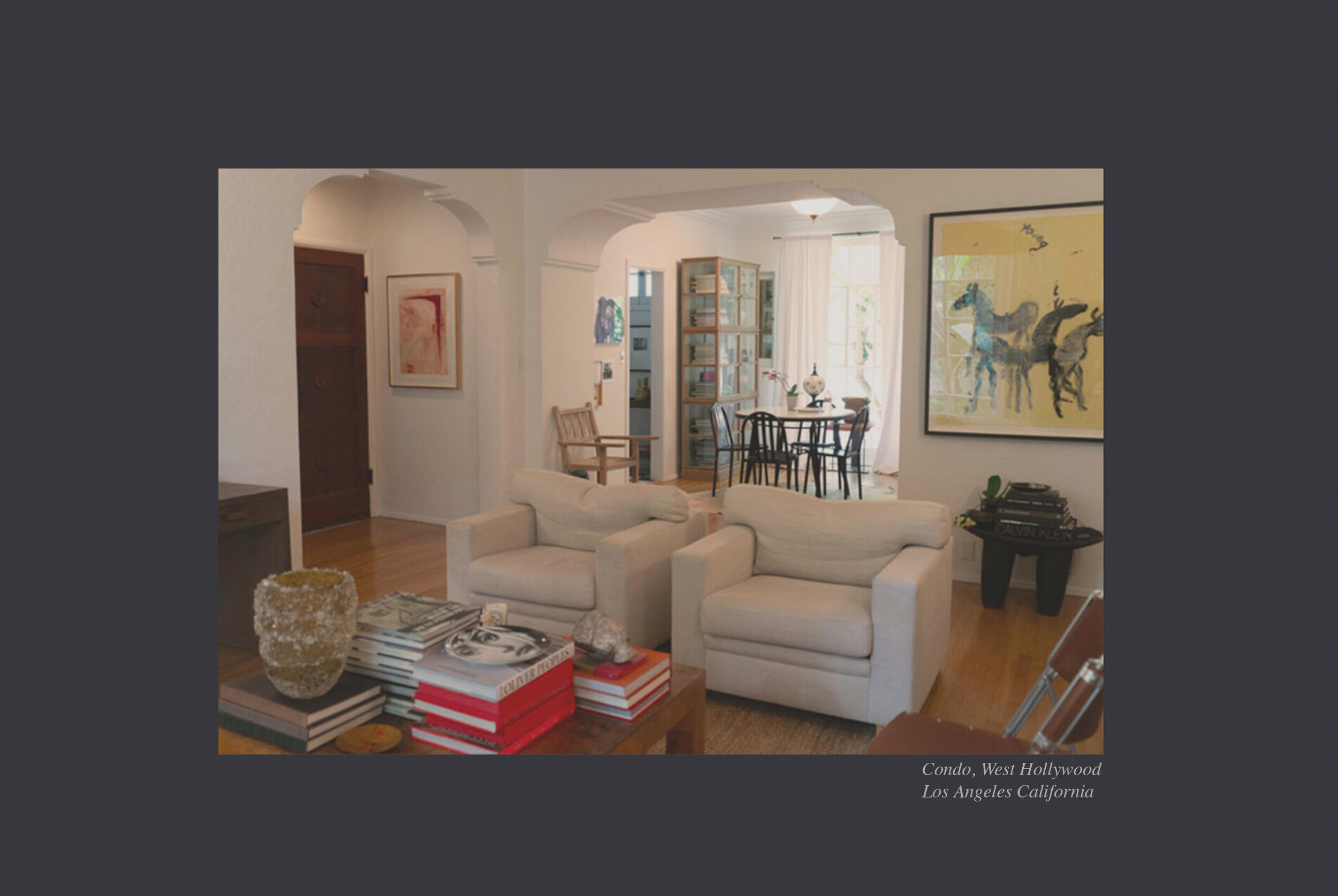 Living room with white sofas, a wooden coffee table with magazines and decor, a round black dining table with chairs, artworks on the walls, a bookshelf, and large windows with white curtains in a condo in West Hollywood, Los Angeles, California.