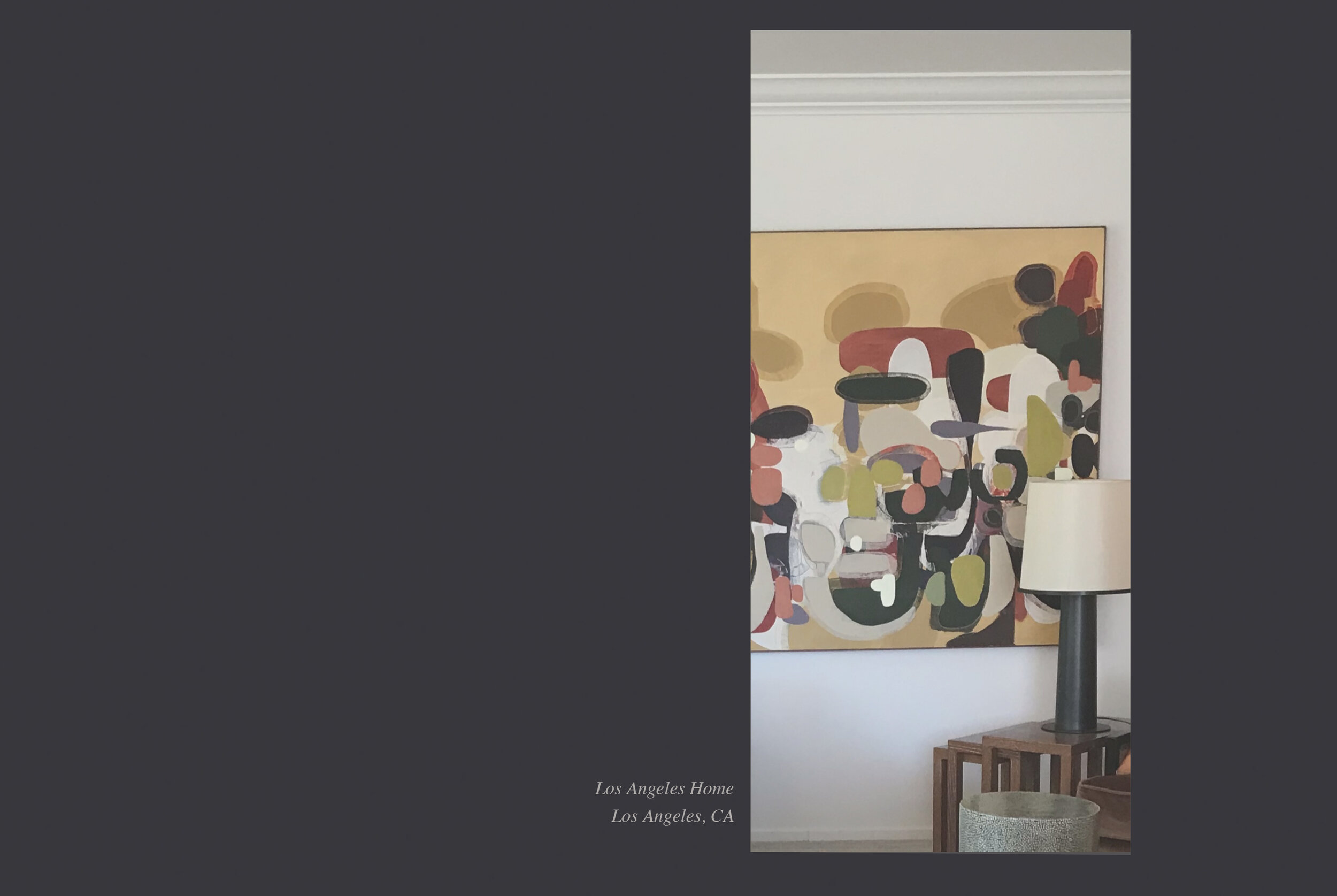 Photograph of the interior of a home with a modern table lamp placed on a wooden table, adjacent to a decorative vase, in front of a white wall with a colorful abstract painting.