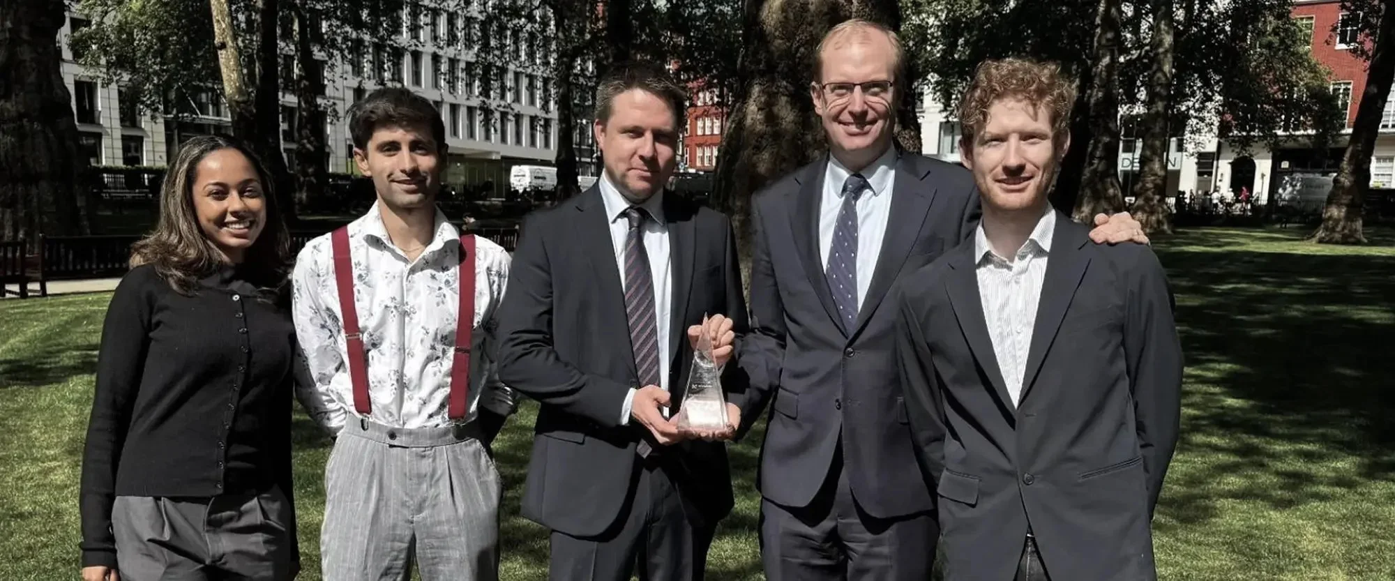 Group of six people in suits and casual clothing standing outside on grass near a large tree, with buildings in the background. Two men in suits are holding an award or trophy.