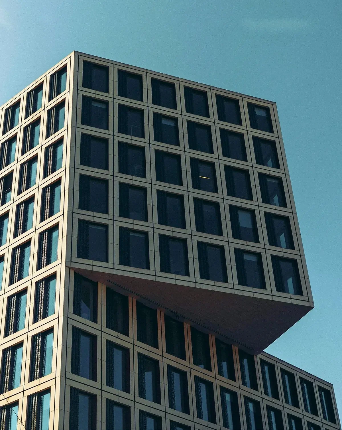 Modern multi-story building with a distinct overhanging section, glass windows, and metal framing, under a clear blue sky.