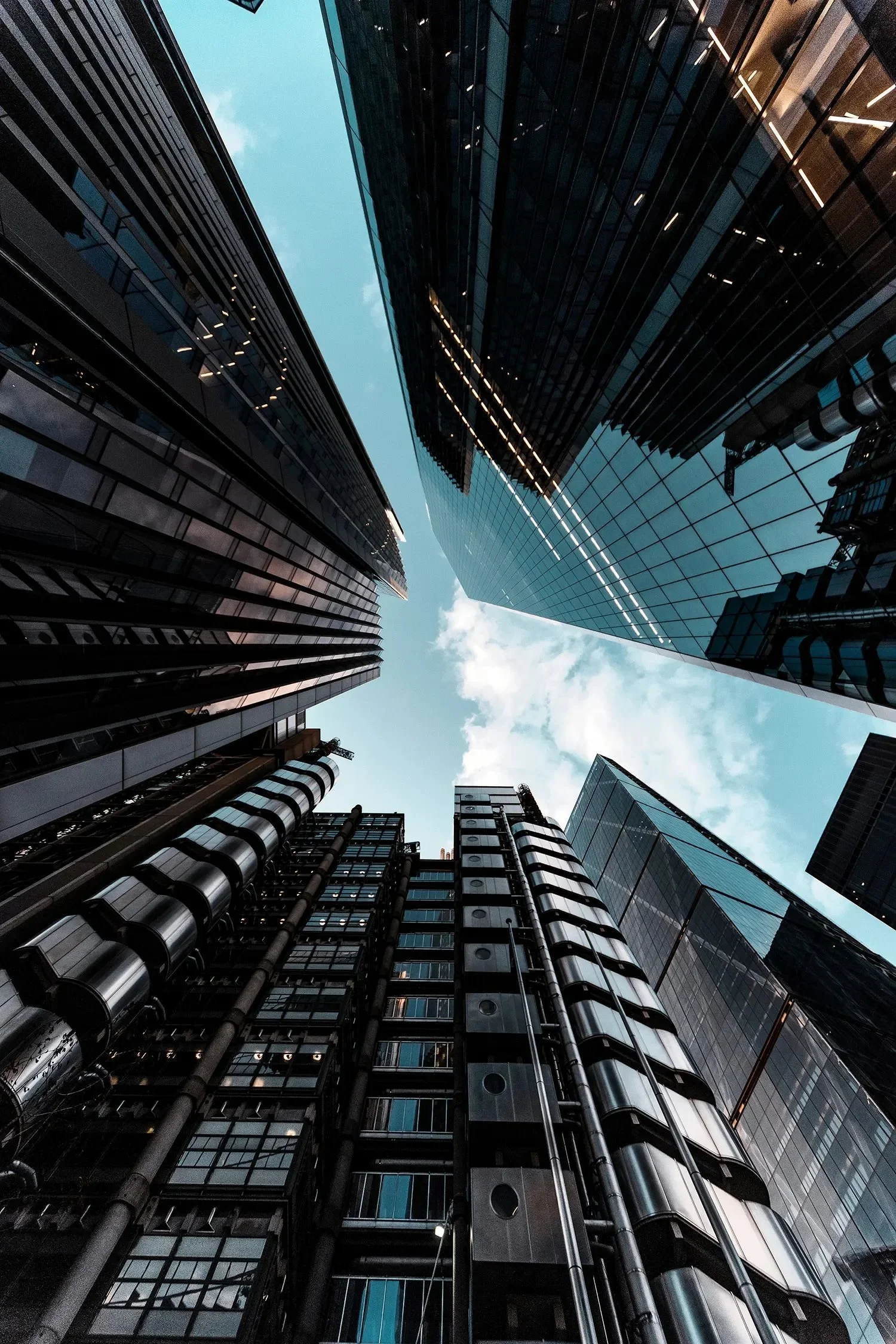 Looking up at tall modern skyscrapers with reflective glass facades in a city, with a blue sky and some clouds above.
