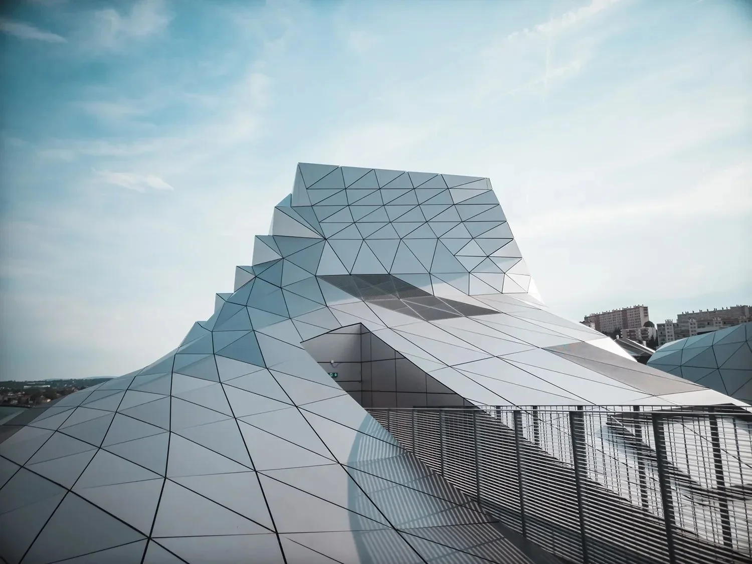 Futuristic building with geometric, angular, metallic panels against a sky with some clouds and city buildings in the background.
