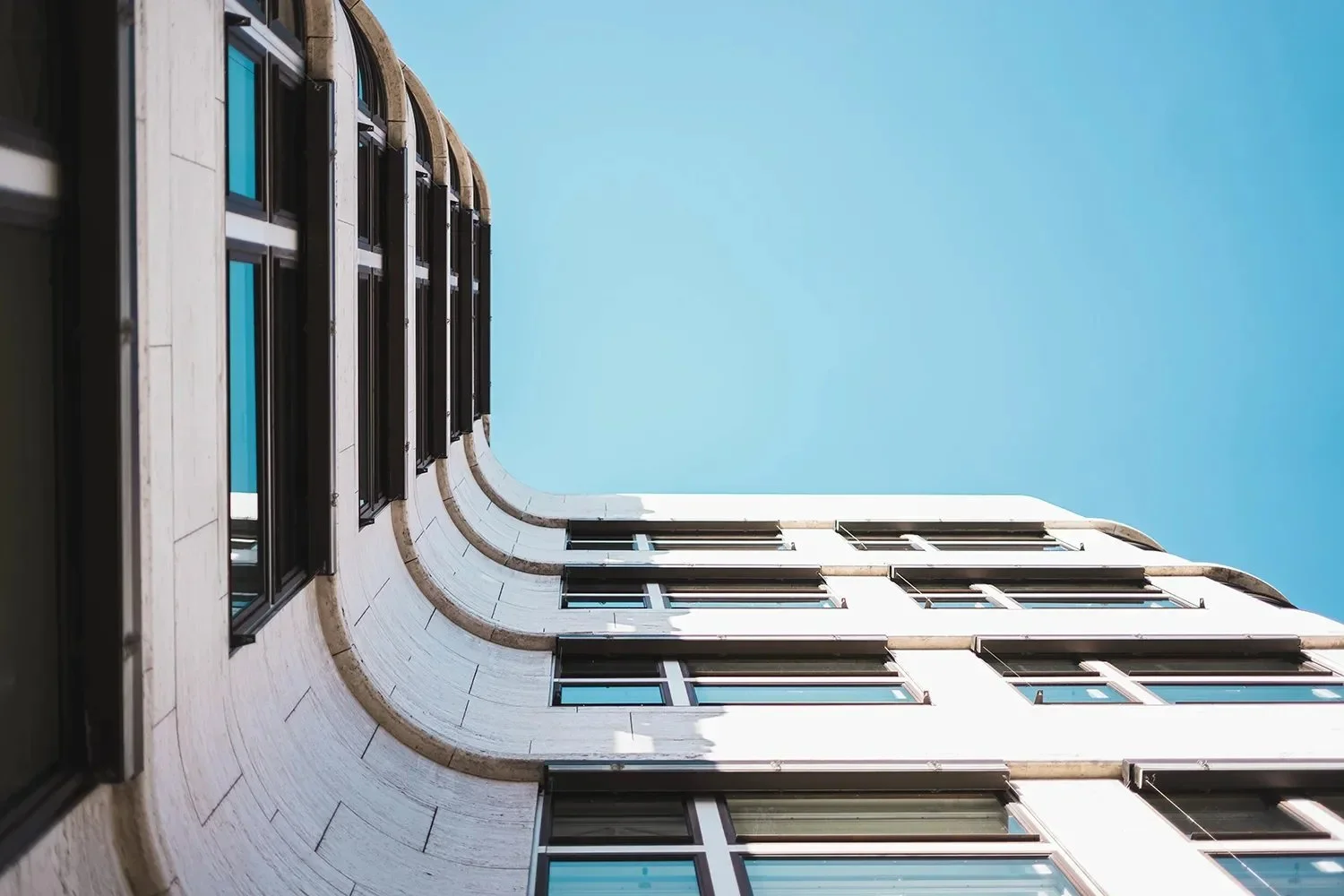Low-angle view of a modern building with white exterior, black window frames, and a curved corner against a clear blue sky.