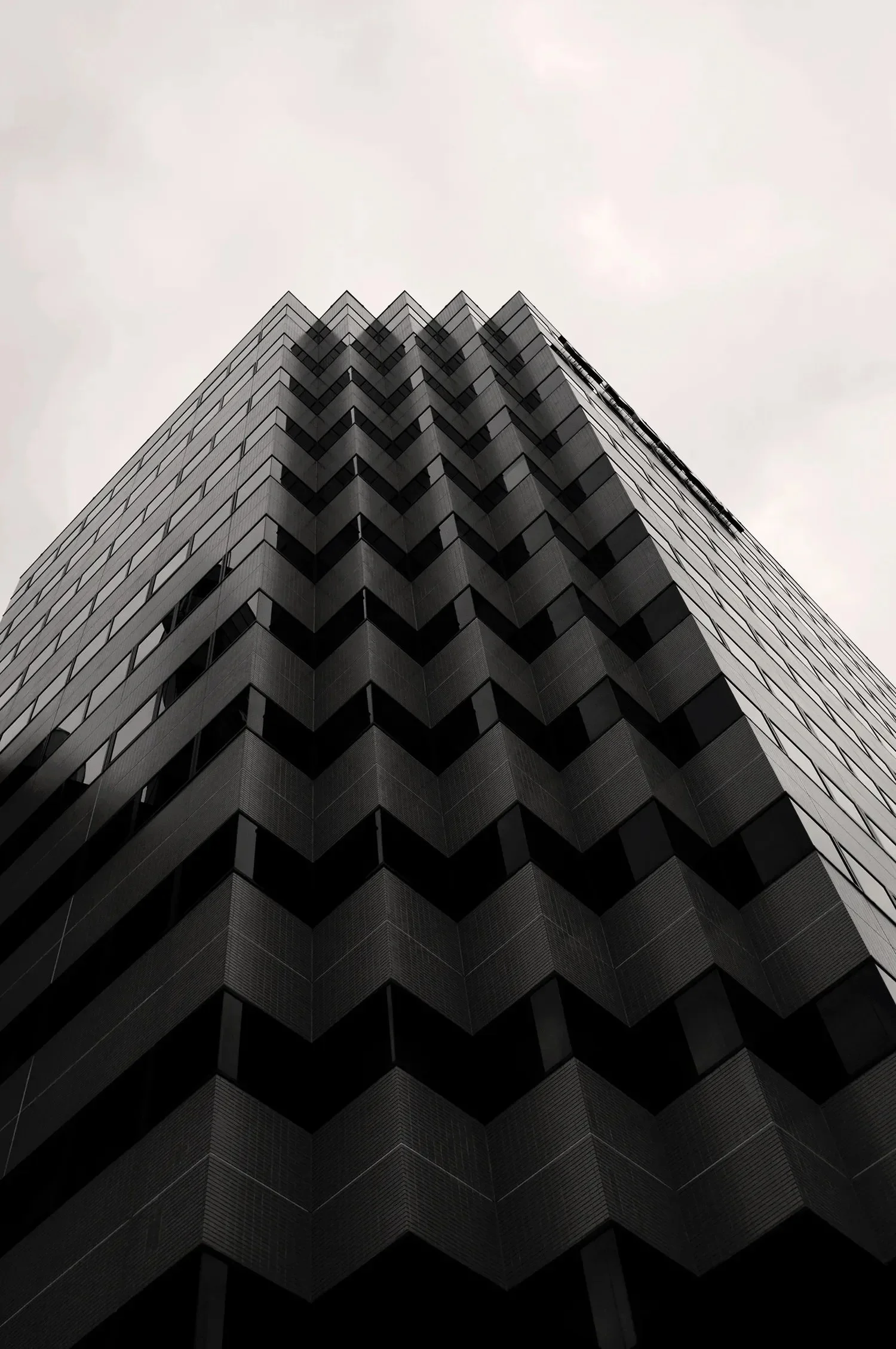 Low-angle view of a tall modern building with a dark facade, featuring a repeated triangular pattern of windows and overhanging balconies, set against a cloudy sky.