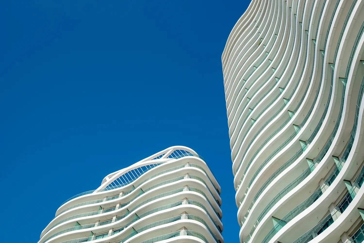 Low-angle view of two modern high-rise buildings with curved white balconies against a clear blue sky.