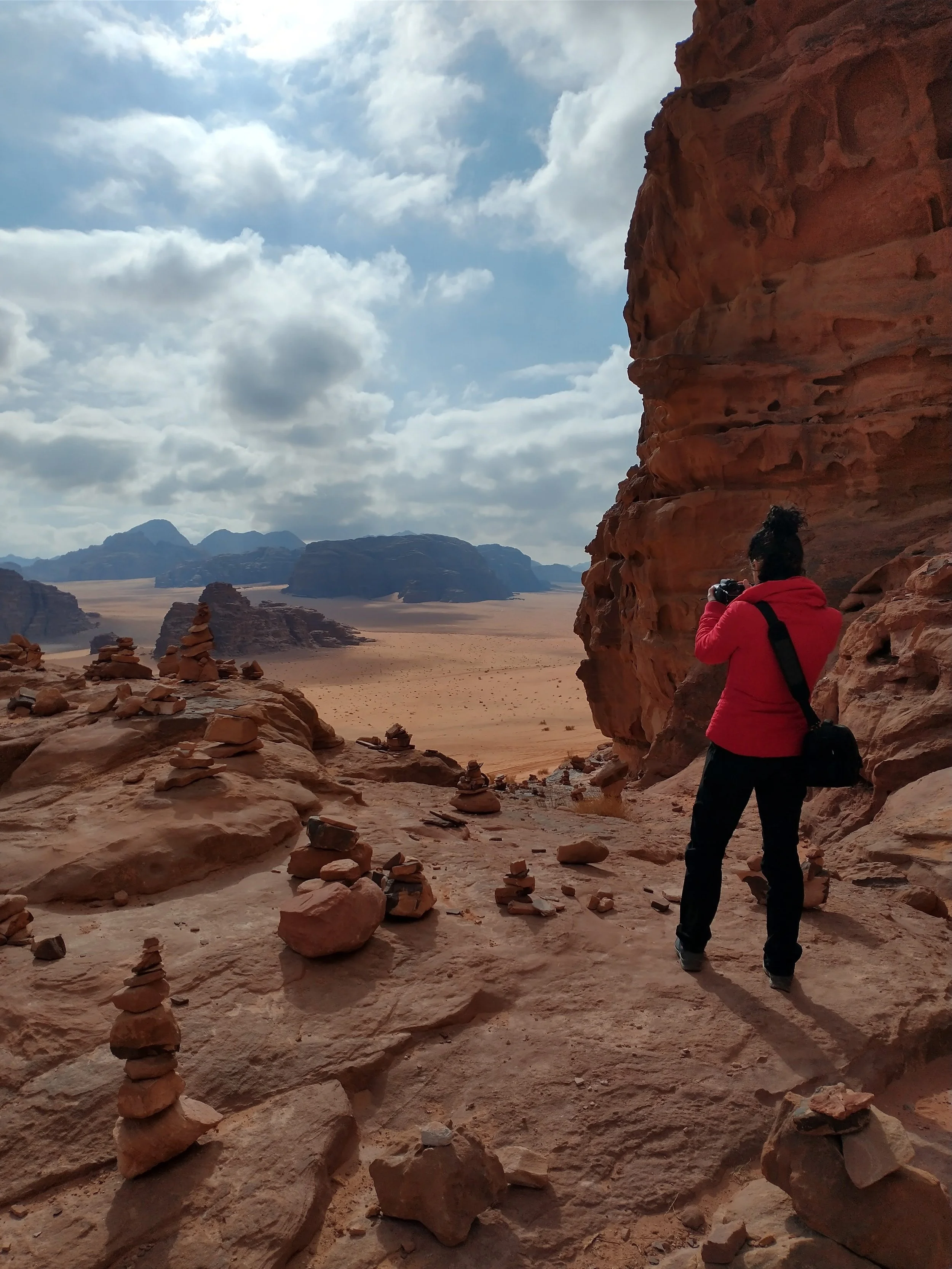 A person in a red jacket taking a photo with a camera in a desert landscape with large rock formations and stacked stones, under a cloudy sky.