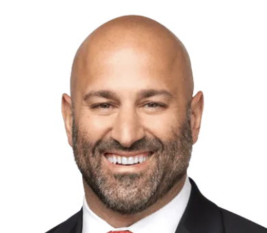 A man in a dark checkered suit with a white shirt smiling against a neutral background.