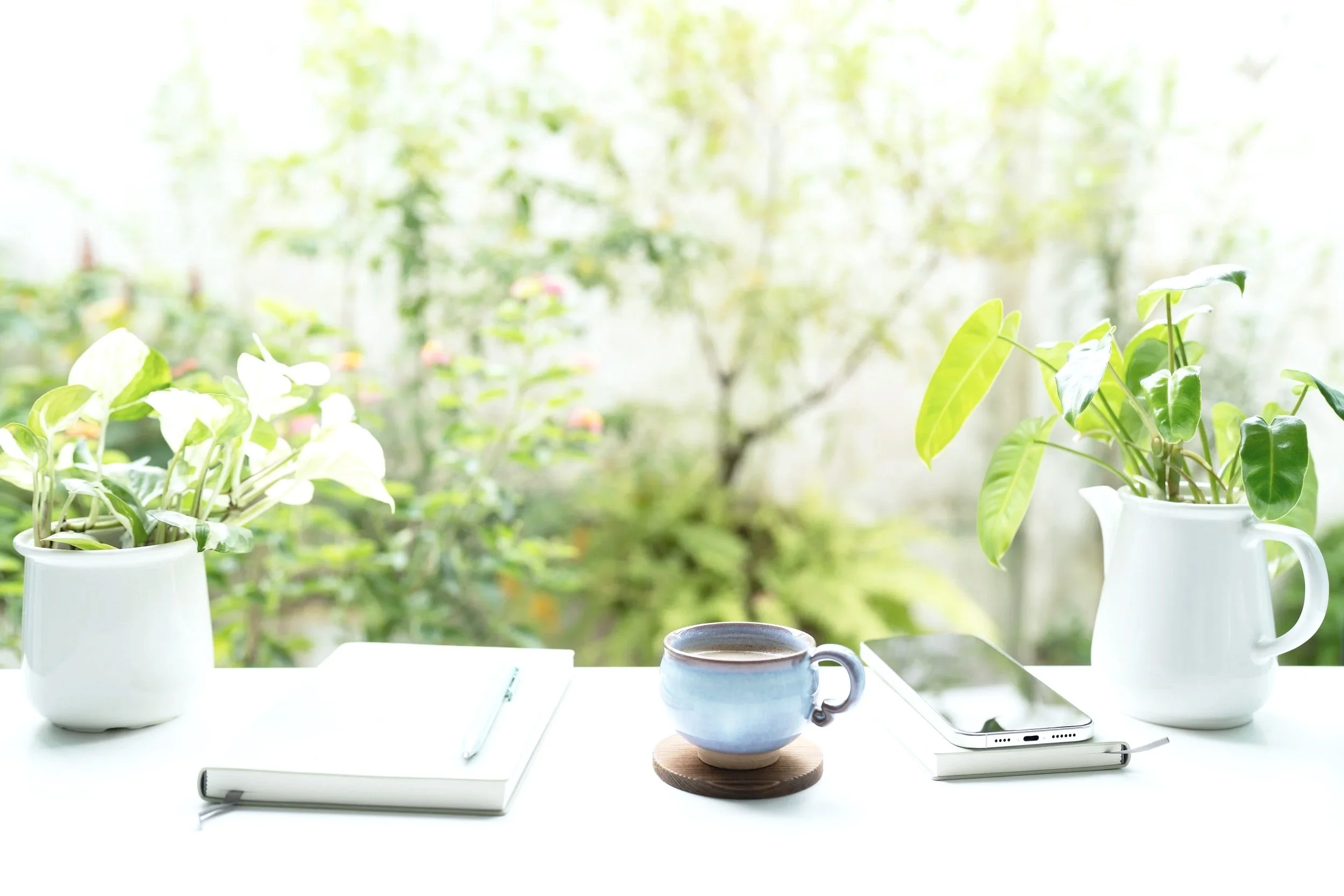 A white table with two potted green plants, a blue coffee mug on a coaster, a smartphone, a notepad with a pen and a white marker, all set in front of a window showing green plants and trees outside.