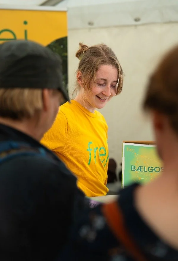 En smilende ung kvinde i en gul t-shirt med teksten "frel" står bag en stand, mens hun snakker med to personer, der er ud af fokus.