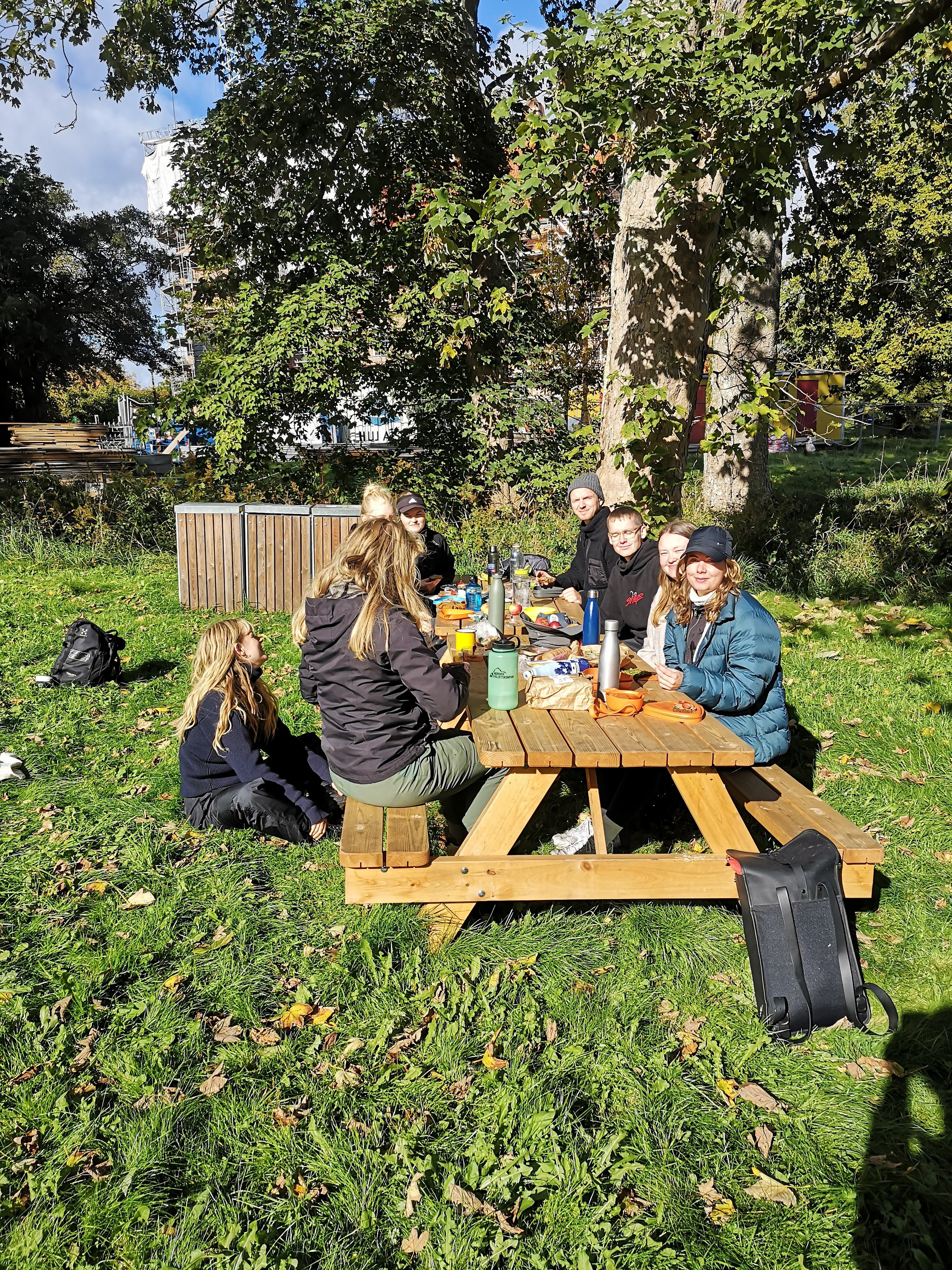 Gruppe mennesker sidder ved picnicbord i en park med træer, sol og græs, og nyder mad udendørs