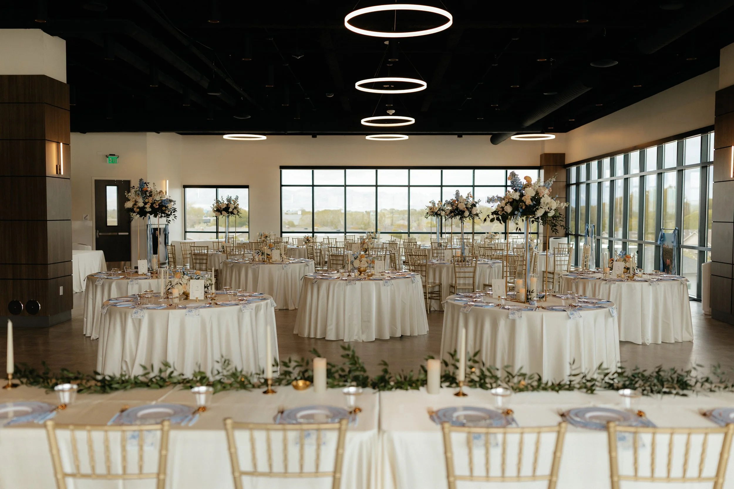 A wedding luncheon setup with a long sweetheart table and several round tables all setup with ivory linens, periwinkle plates, and tall flower arrangements.