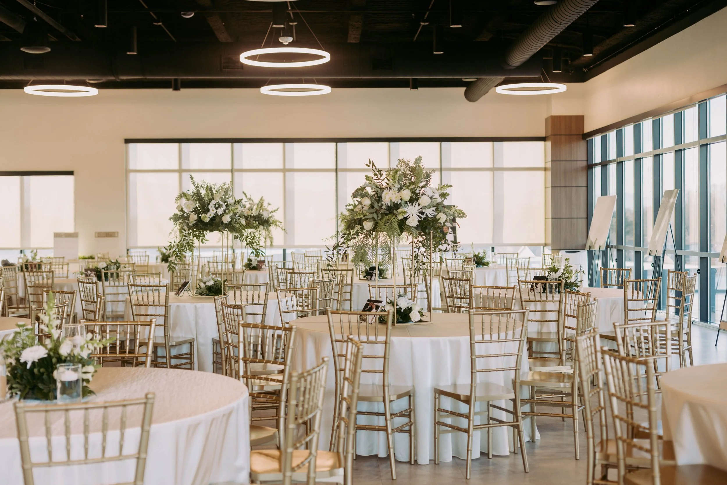 Round tables setup with white linens, surrounded by gold chairs, and topped with tall white and green floral centerpieces.
