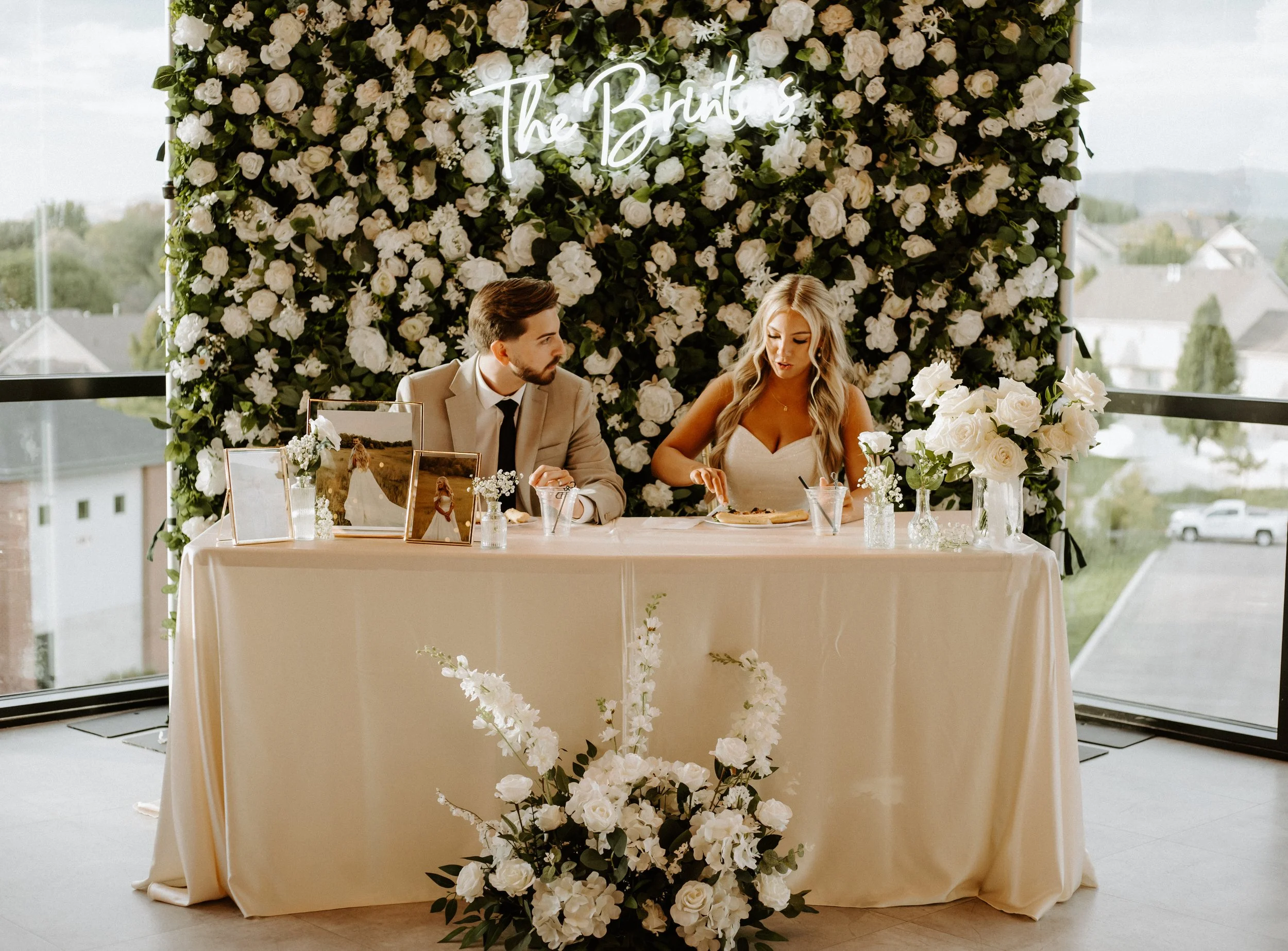 Bride and groom sit at the sweetheart table that's adorned with white florals and lush greenery.