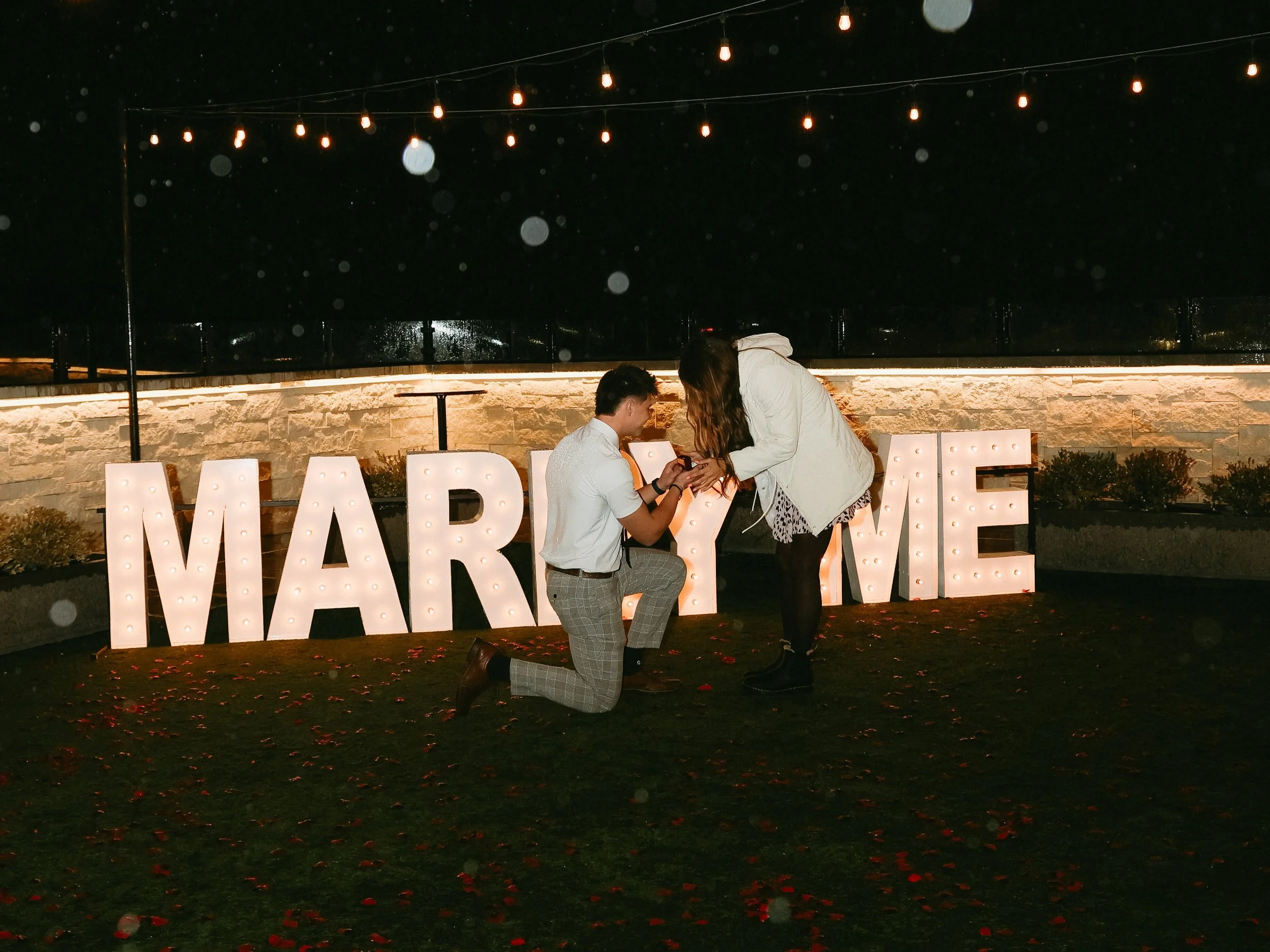 Man is on one knee proposing to a woman in front of LED letters that say marry me.