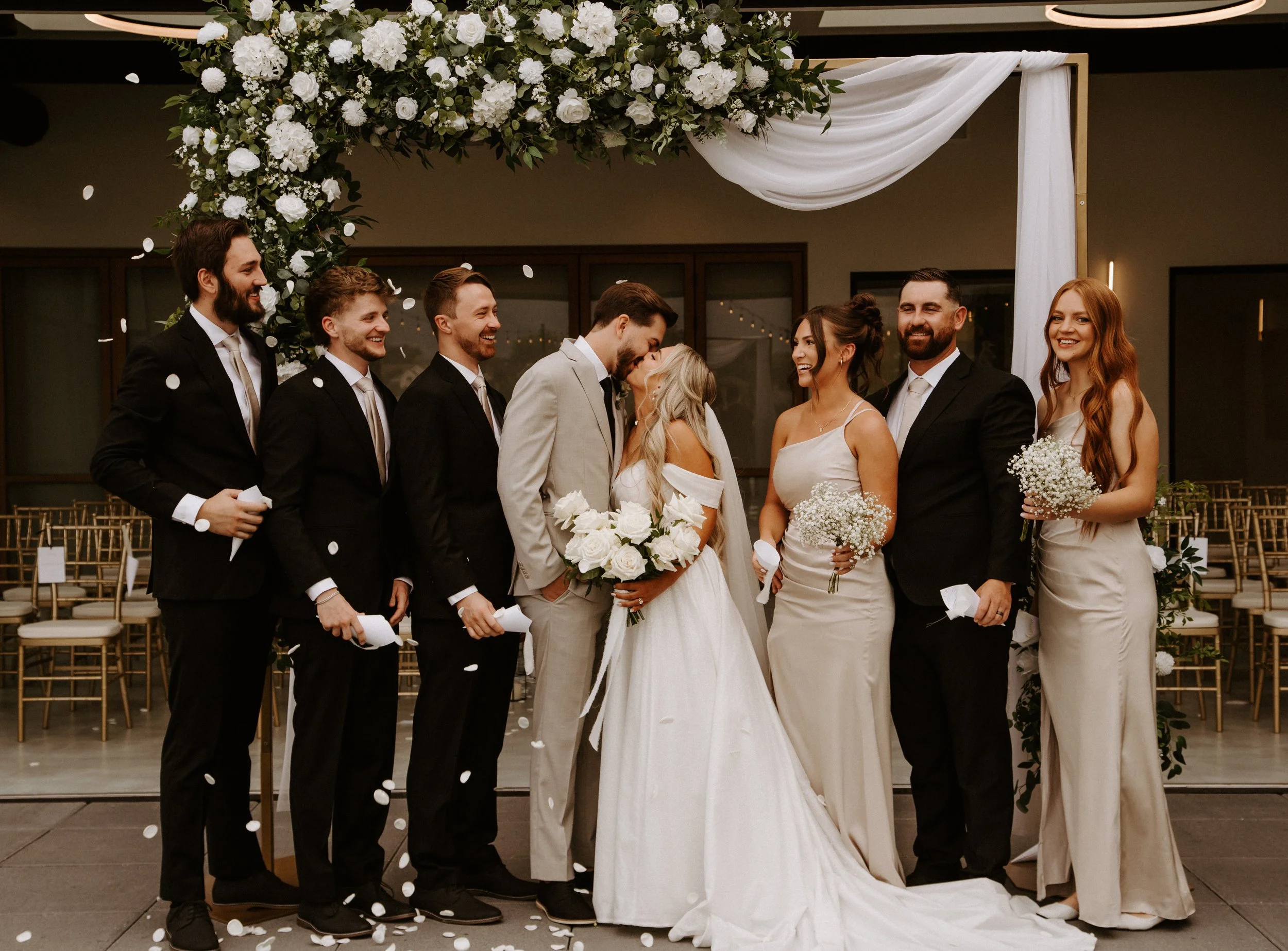 The bride and groom kiss in the center of the bridal party under a square wedding arch.
