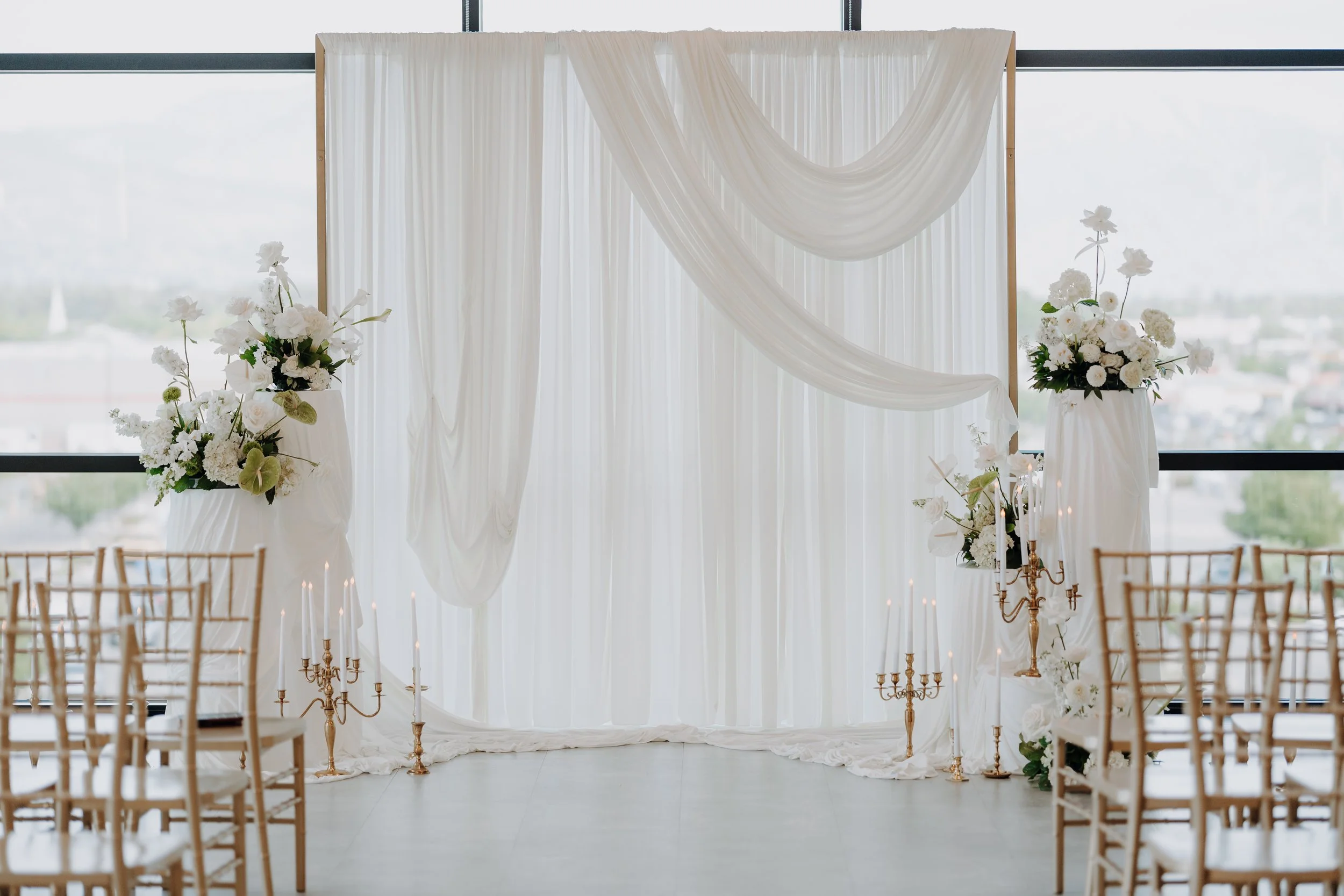 Wedding ceremony backdrop with white draping, white florals, and gold candle sticks.