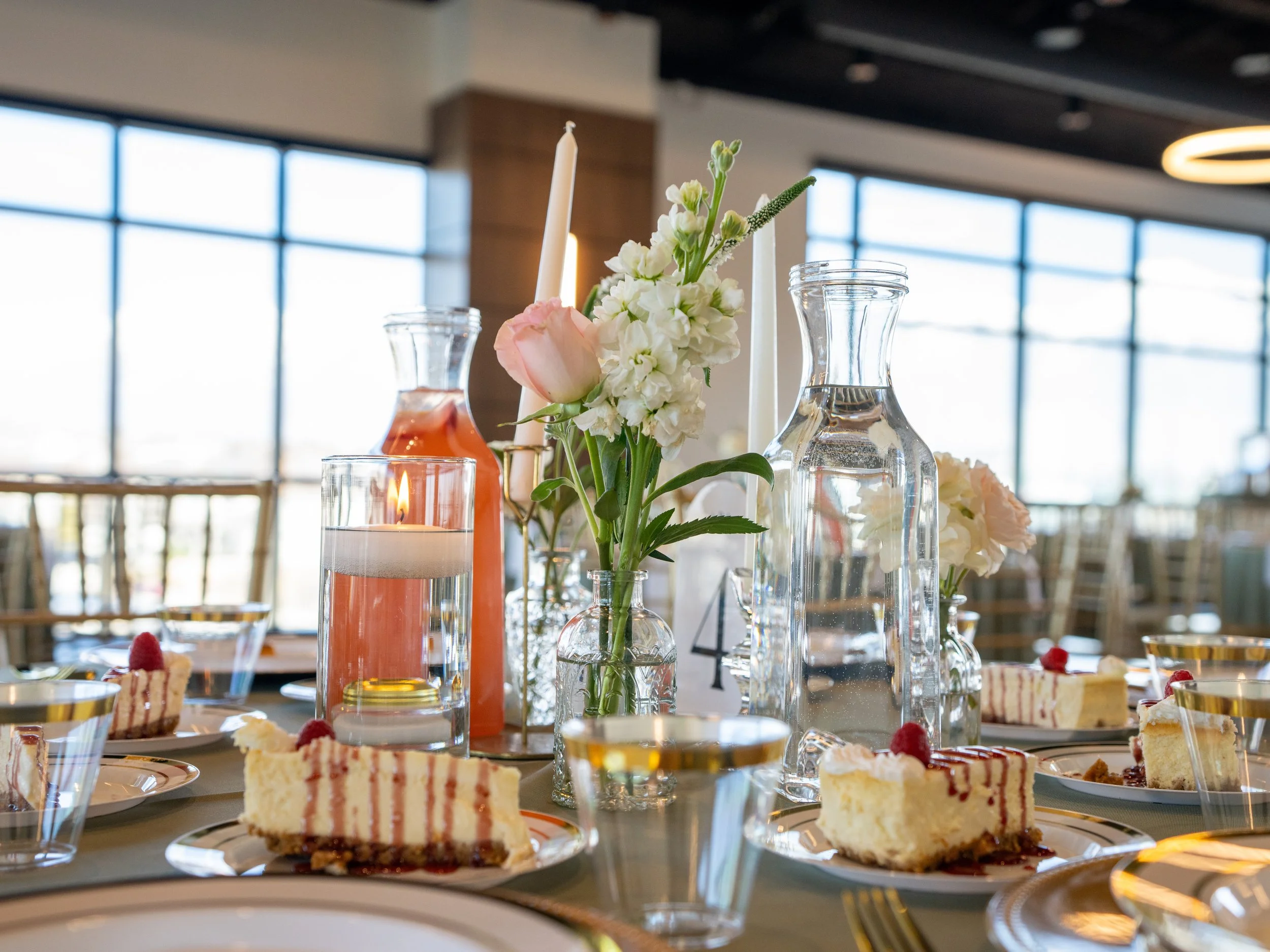 Wedding table set up with sage green linens, clear cups, pink and white floral centerpieces, and a slice of cheesecake at each seat.