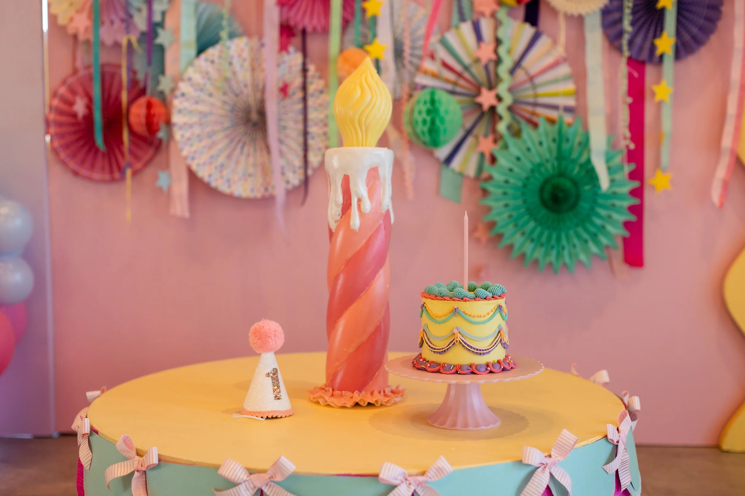 Yellow birthday cake next to a giant coral faux birthday candle and little white party hat.