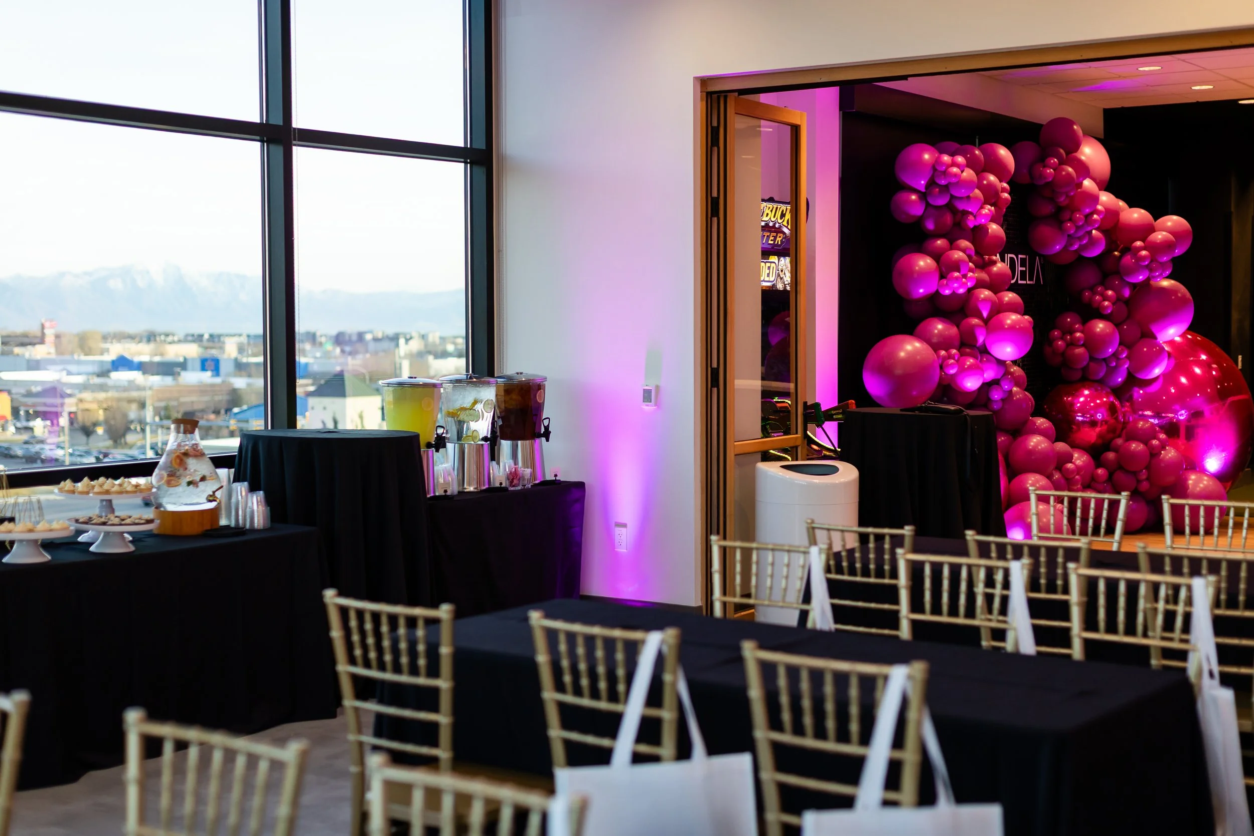 Corporate event setup using rectangle tables with black linens, gold chairs, and a large pink balloon backdrop.