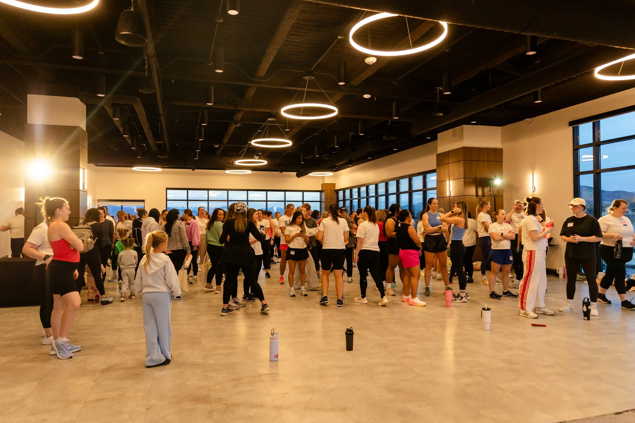A large group of women is standing in an indoor venue wearing exercise clothing.