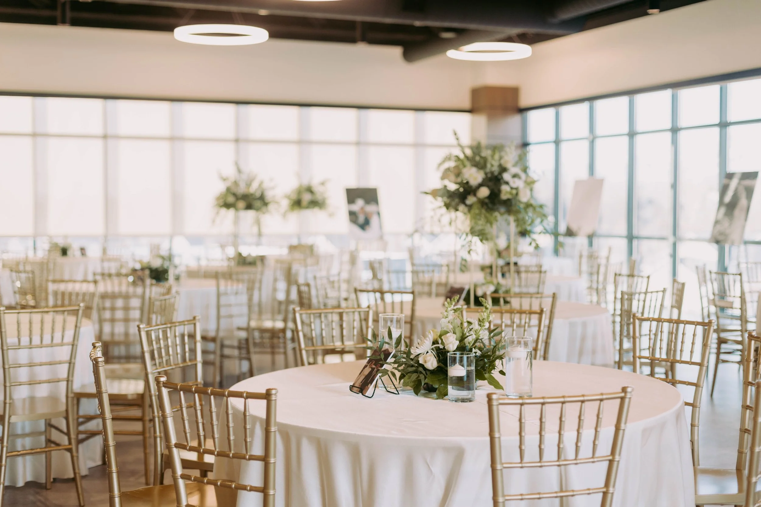 Round tables setup in an indoor venue, draped with white linens and topped with green and white floral arrangements.