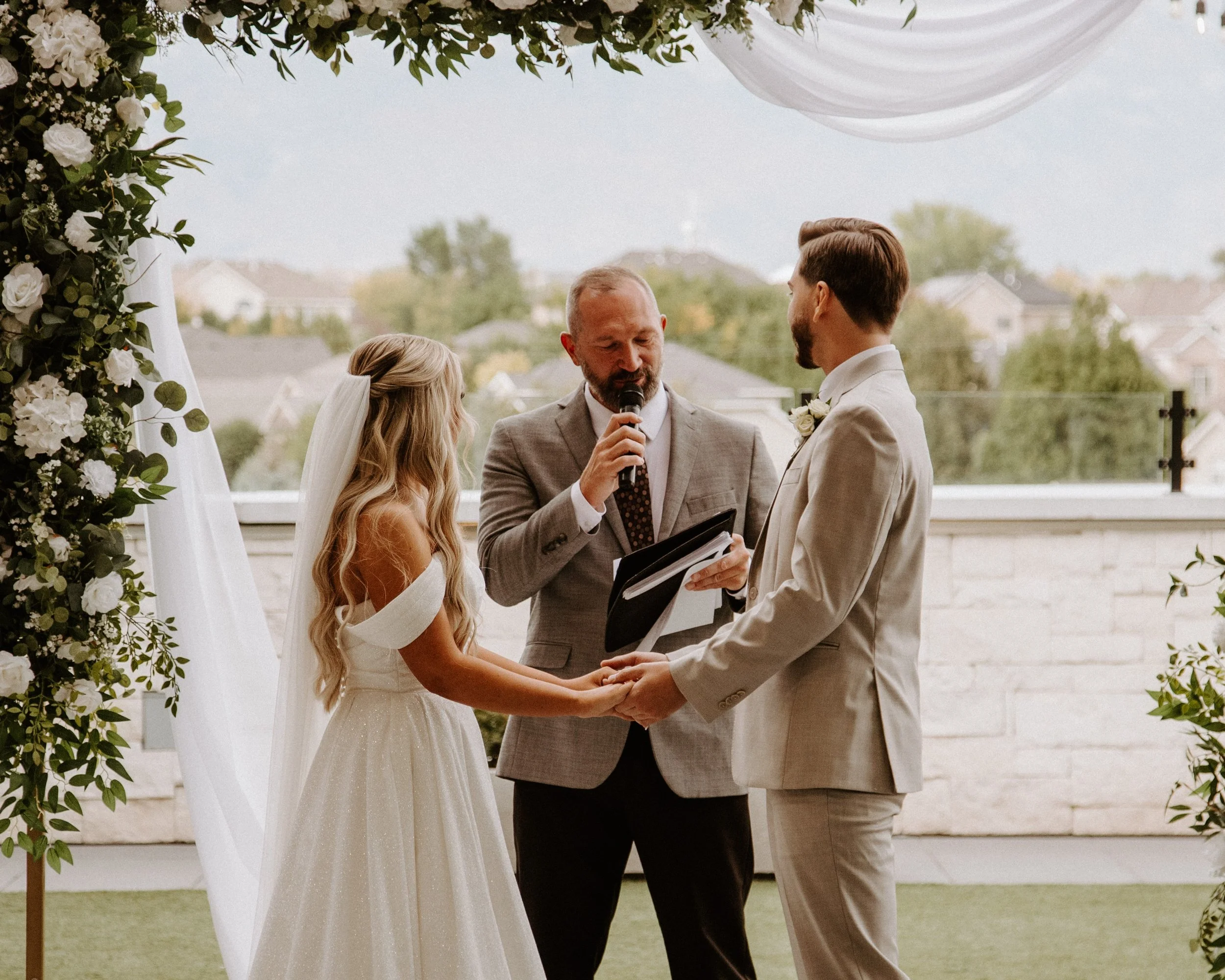 Bride and groom stand hand-in-hand while looking at the wedding officiant.
