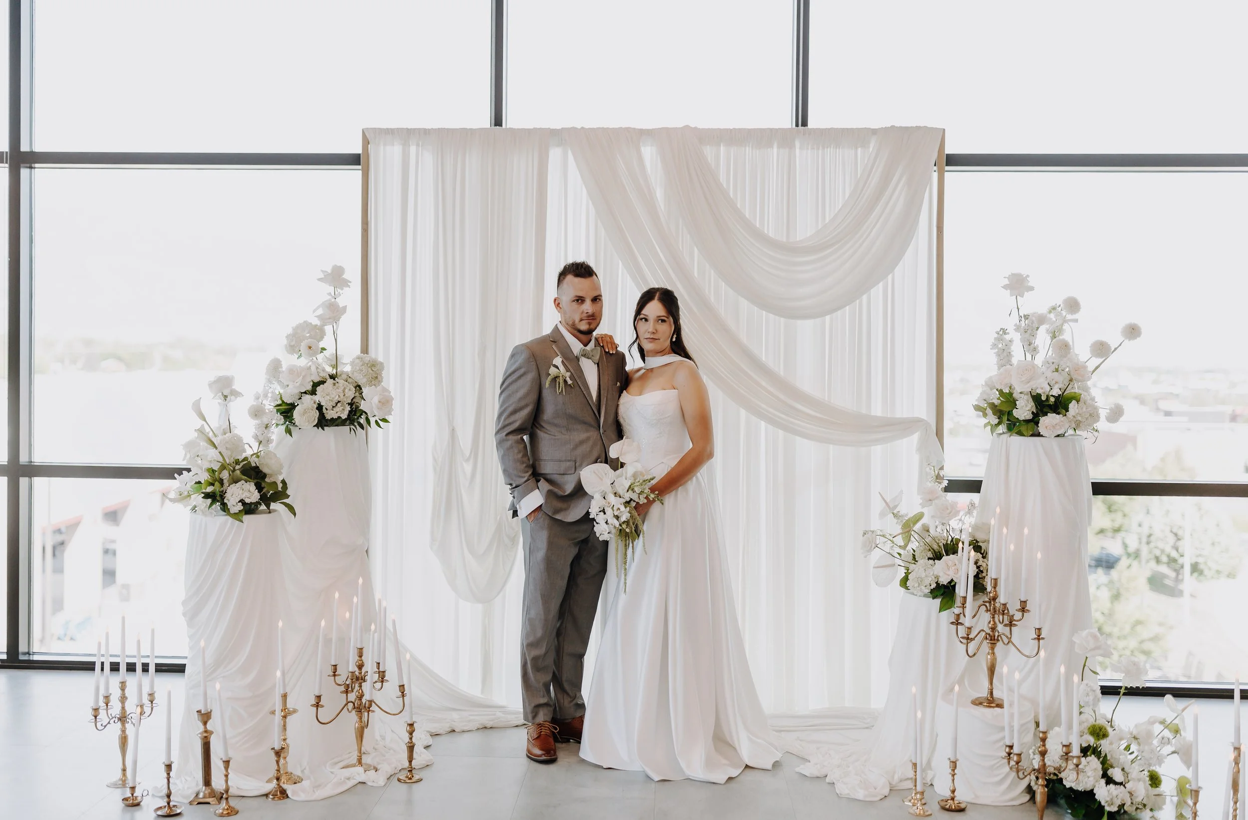 The bride and groom post in front of a white draped backdrop with white flowers and gold candlesticks around them.